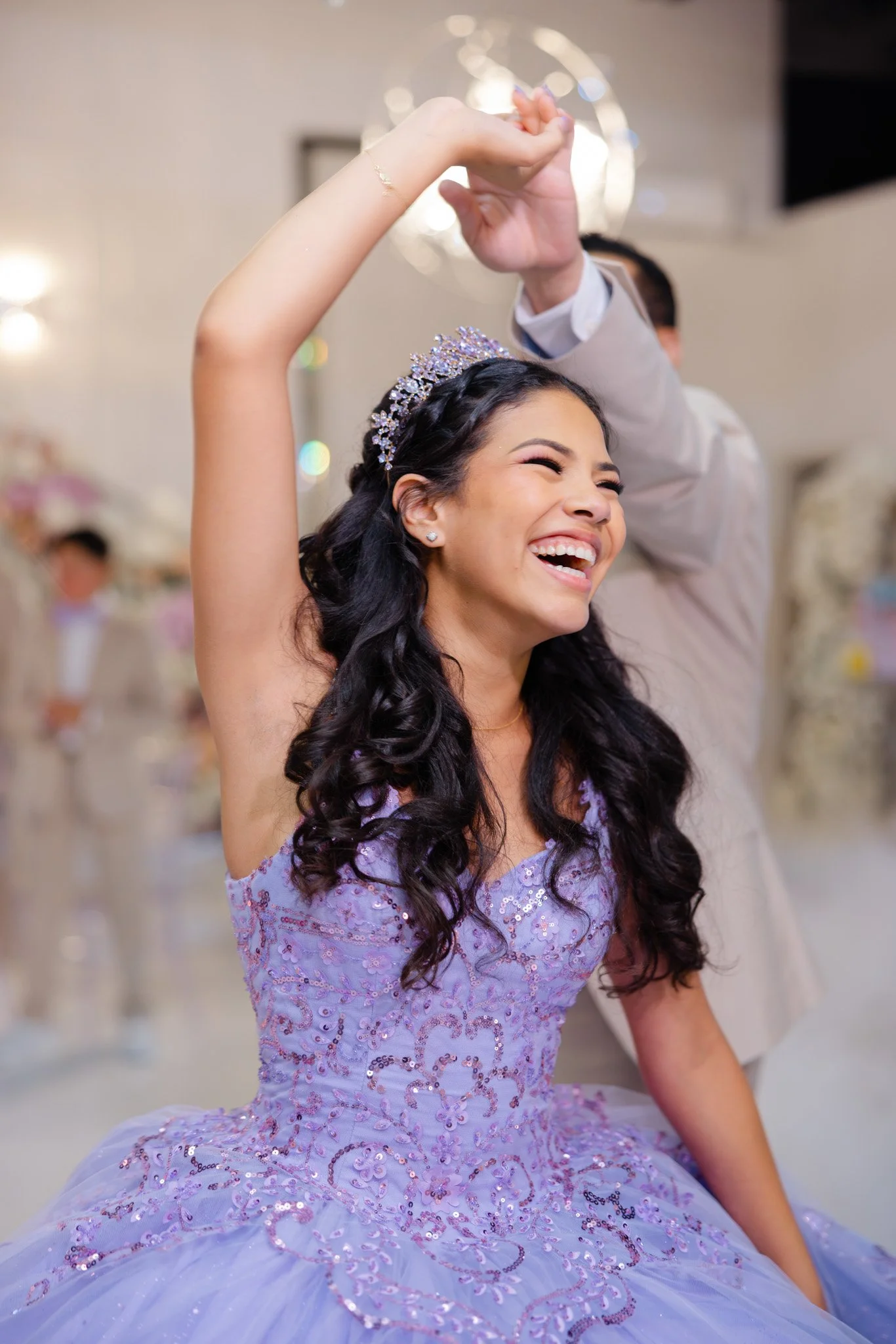 A young woman in a lilac, sequined ball gown with an off-shoulder neckline, smiling joyfully and wearing a tiara, at a celebration or quinceañera.