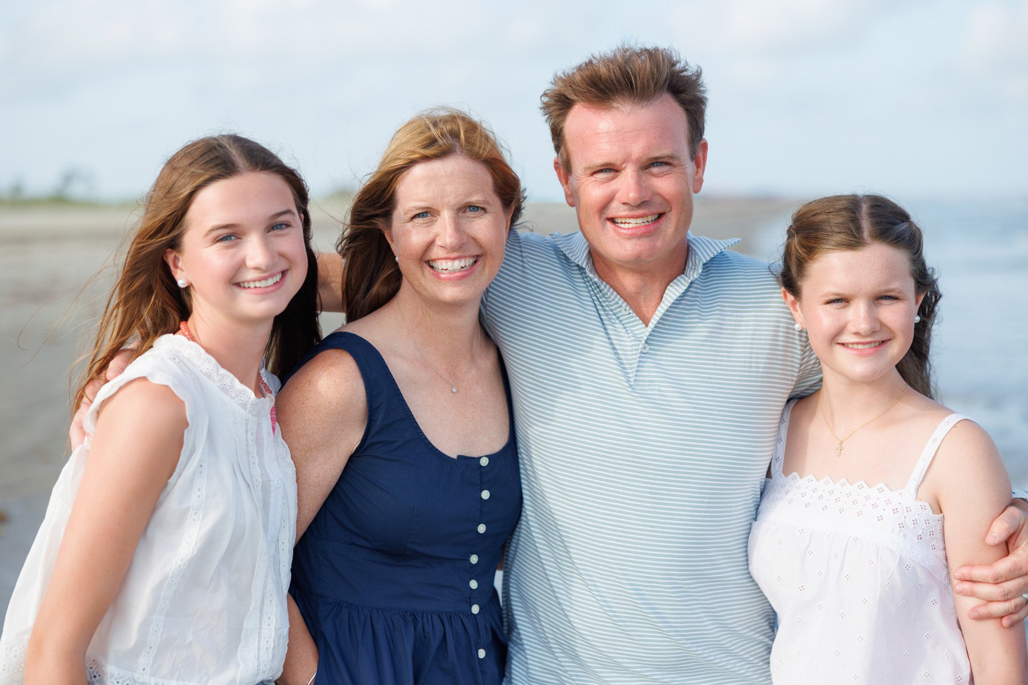 A family of four smiling outdoors at the beach with light clouds in the sky. They are standing close together, arms around each other.