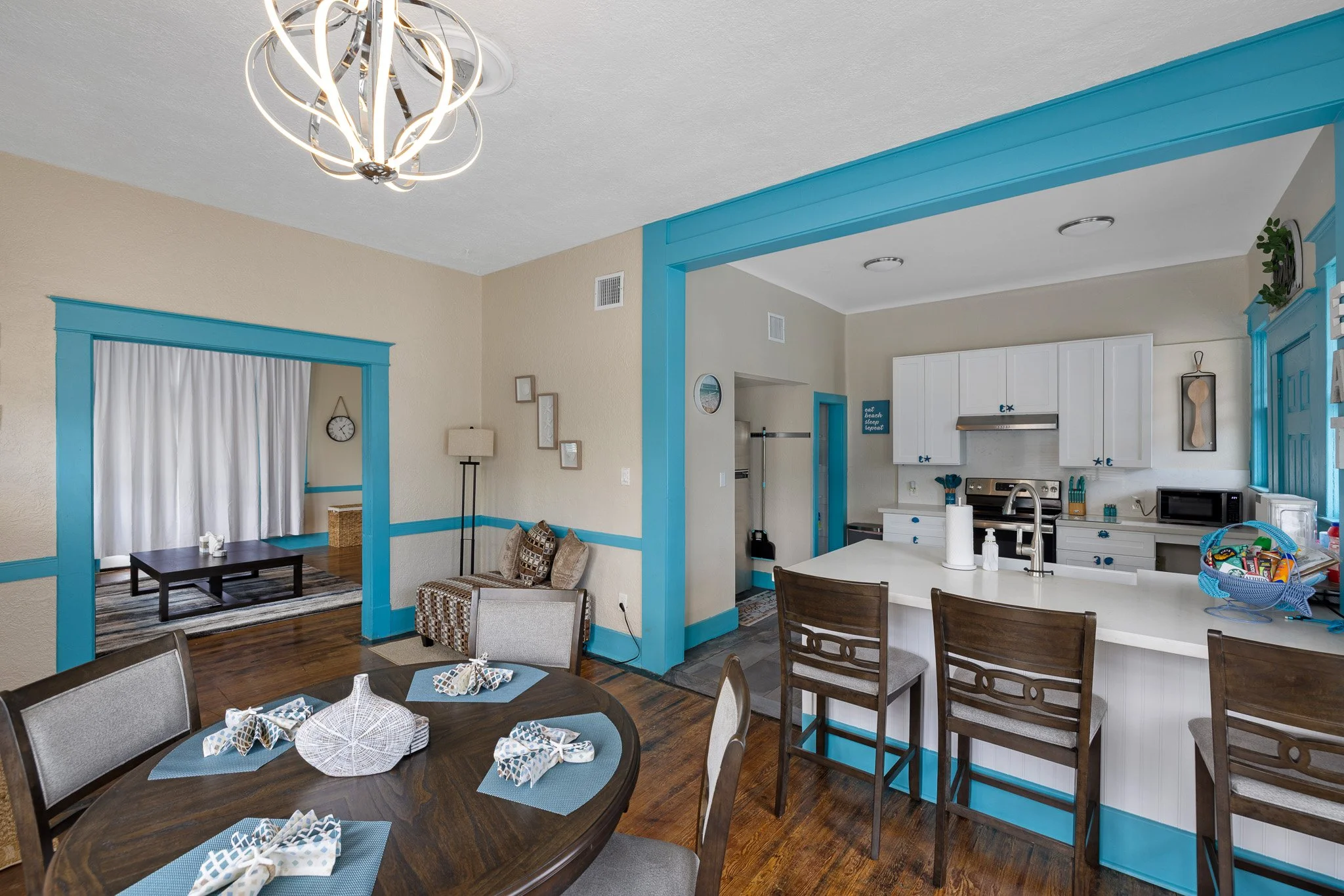 View of a dining area and kitchen in a house, with blue trim on walls and furniture, a wooden dining table set with placemats and napkins, a kitchen island with chairs, white cabinets with blue accents, stainless steel appliances, and a small living
