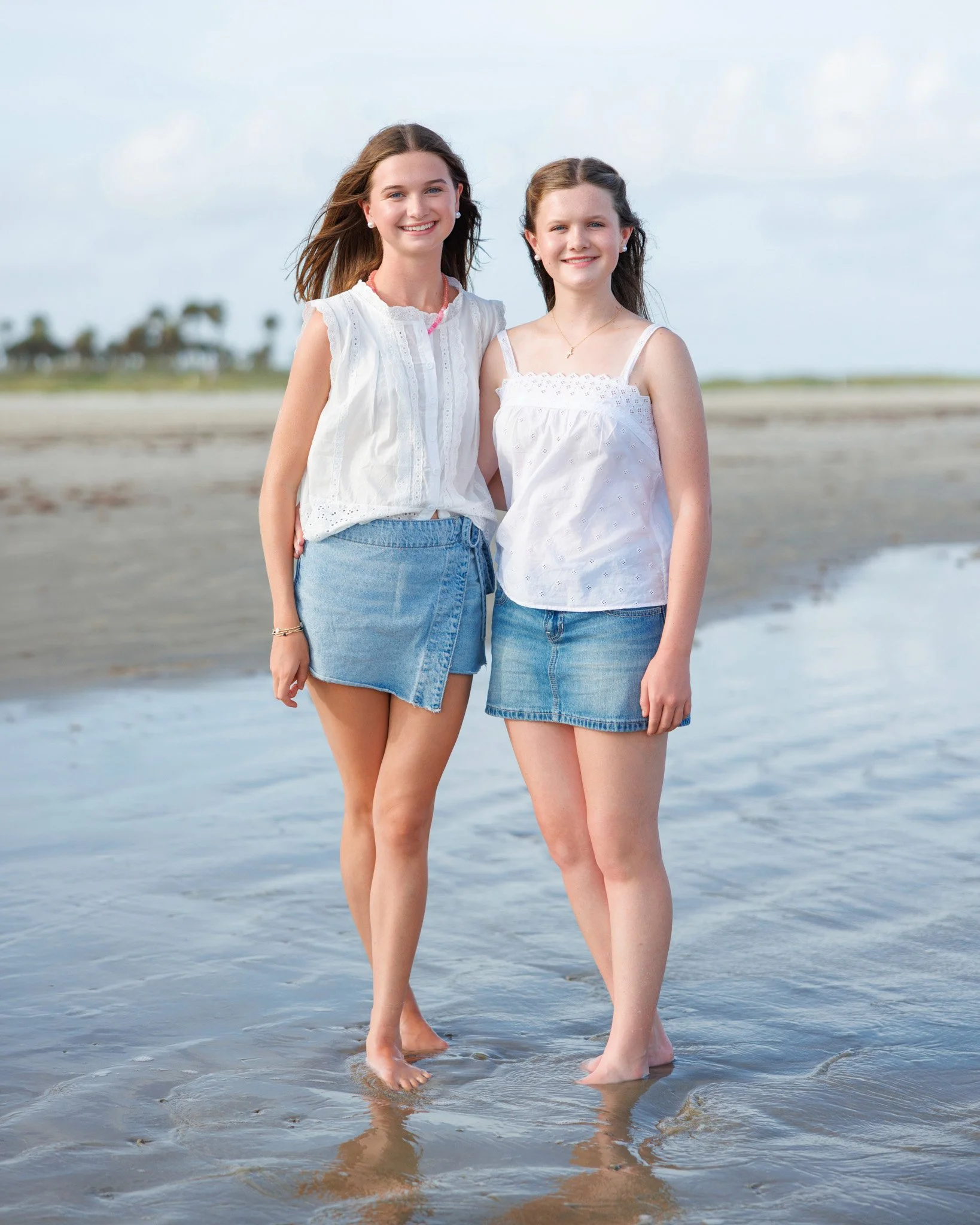 Two young women in white tops and denim skirts standing on a sandy beach with water at their feet, smiling at the camera.