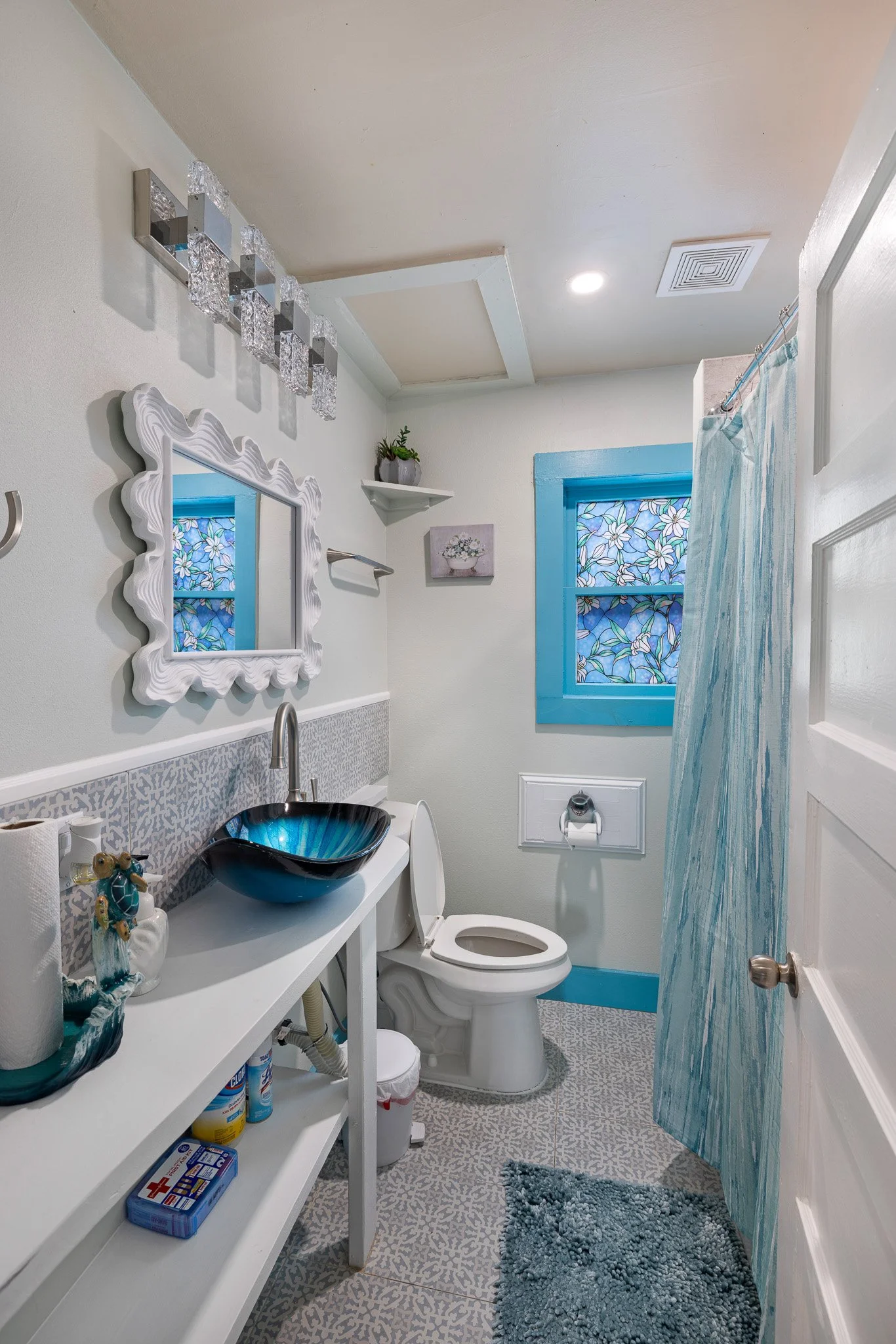 Bathroom with a white vanity, black glass sink, and blue accents including a window, trim, and shower curtain, decorated with stained glass art and small plants.