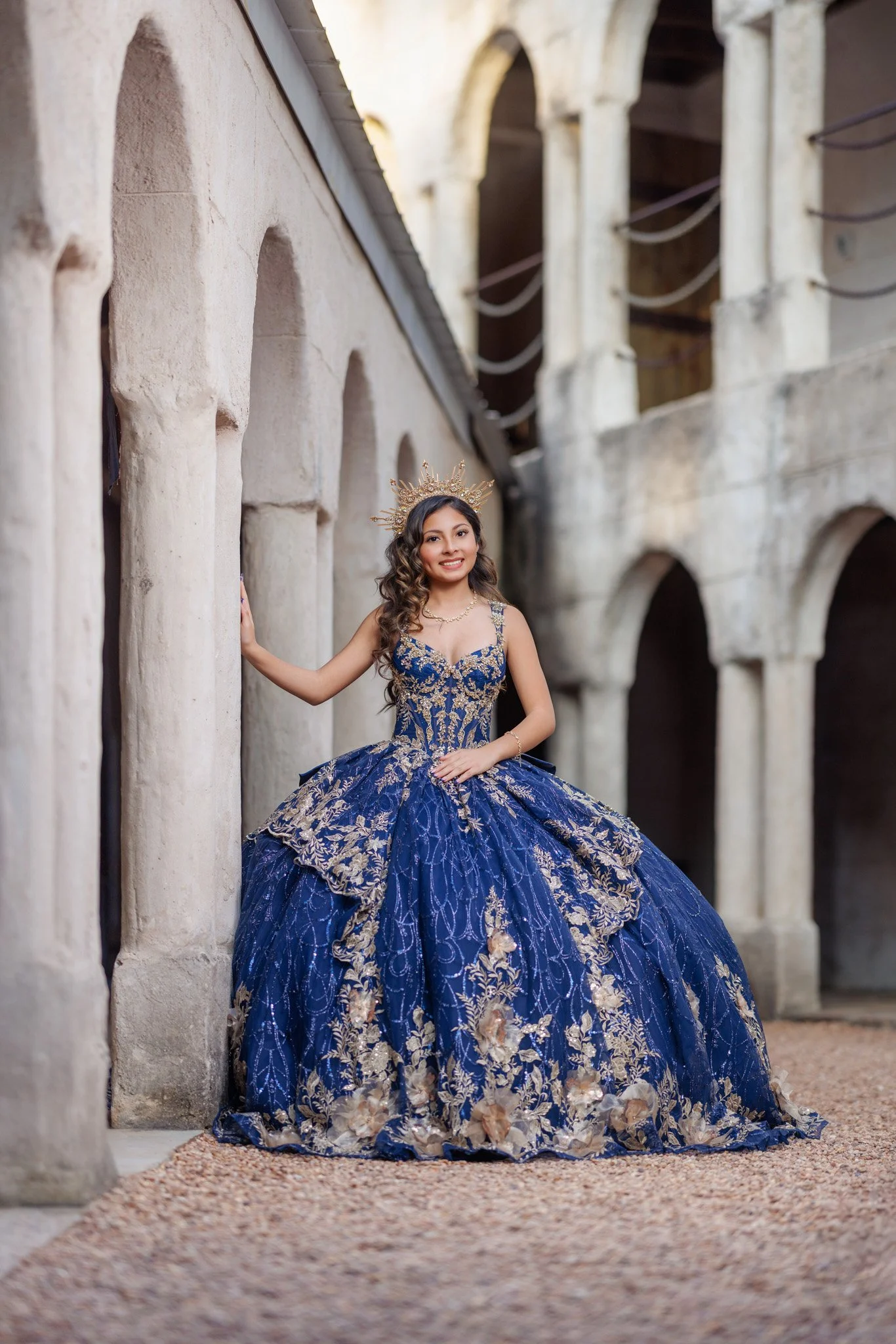 A young woman dressed in a royal blue gown with intricate gold embroidery and a full skirt, standing in a historic stone building with arches. She wears a golden crown and has long, wavy hair, smiling at the camera.