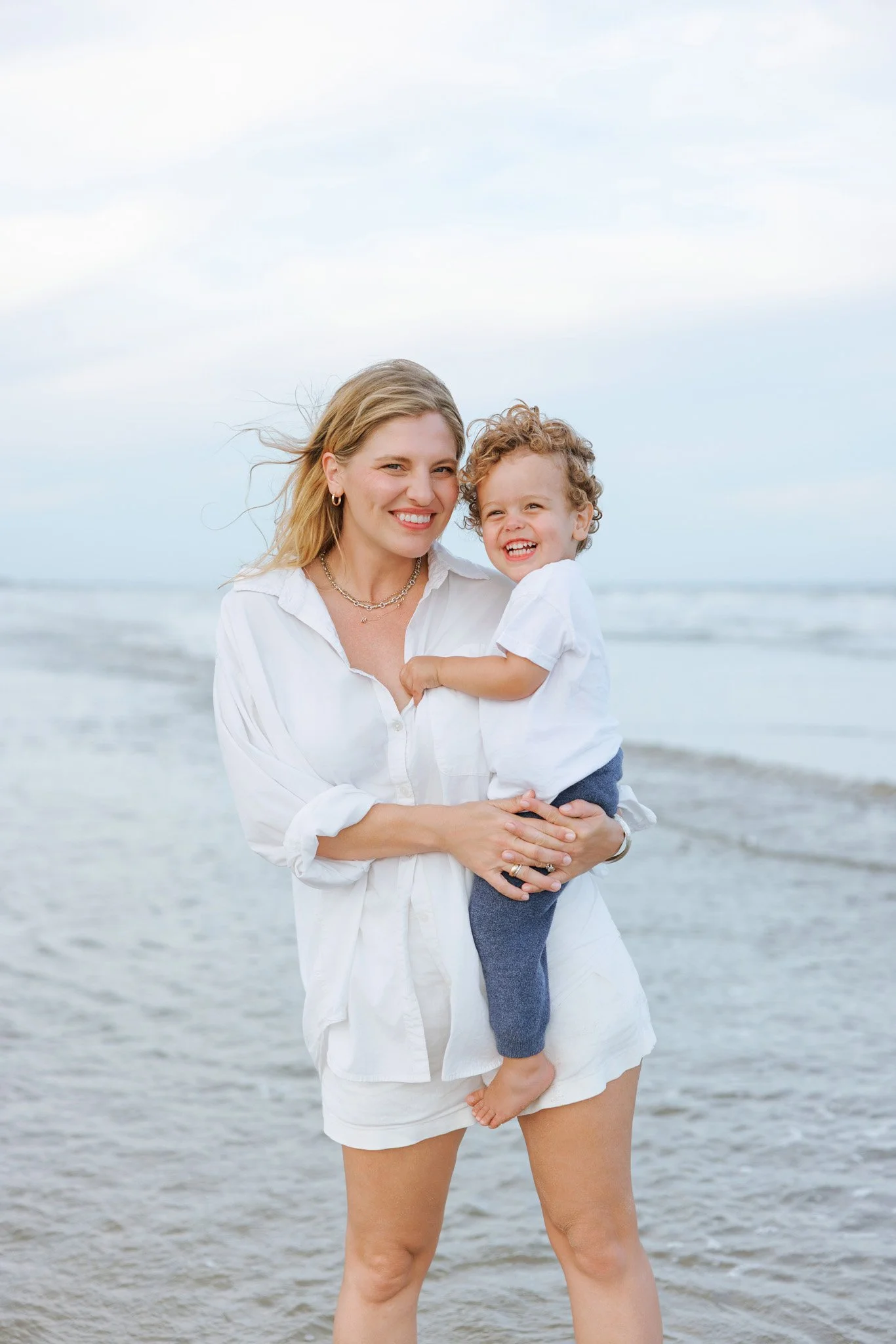 A woman holding a young boy while standing on the beach with the ocean in the background, both smiling.