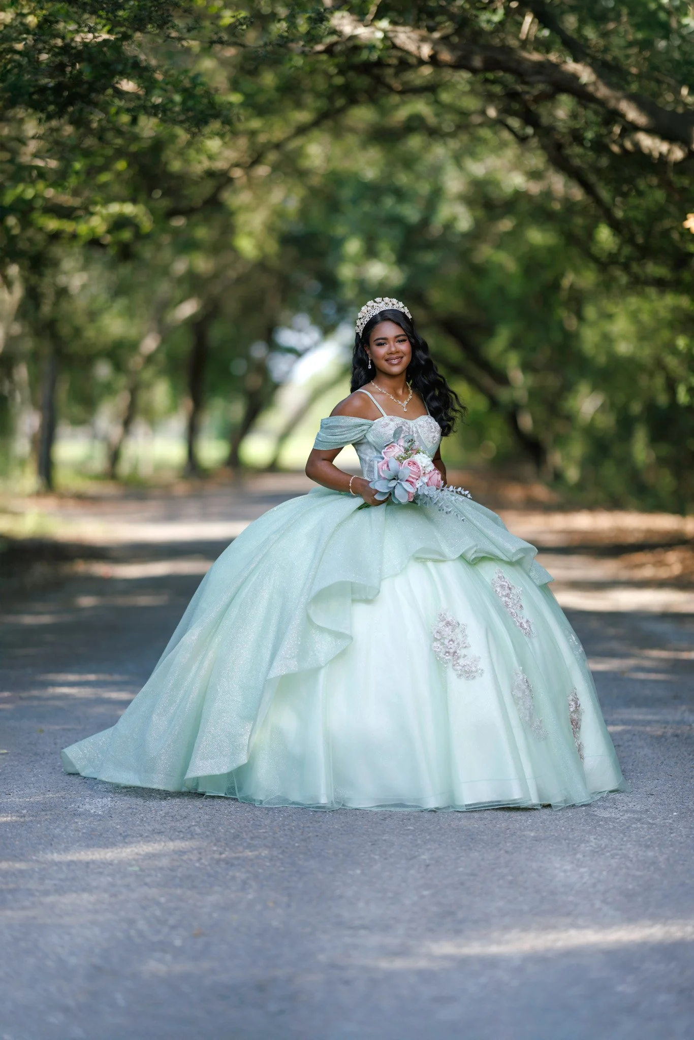 A young woman in an elegant green ball gown and floral tiara posing outdoors on a tree-lined path, holding a bouquet of pink roses and wearing jewelry.