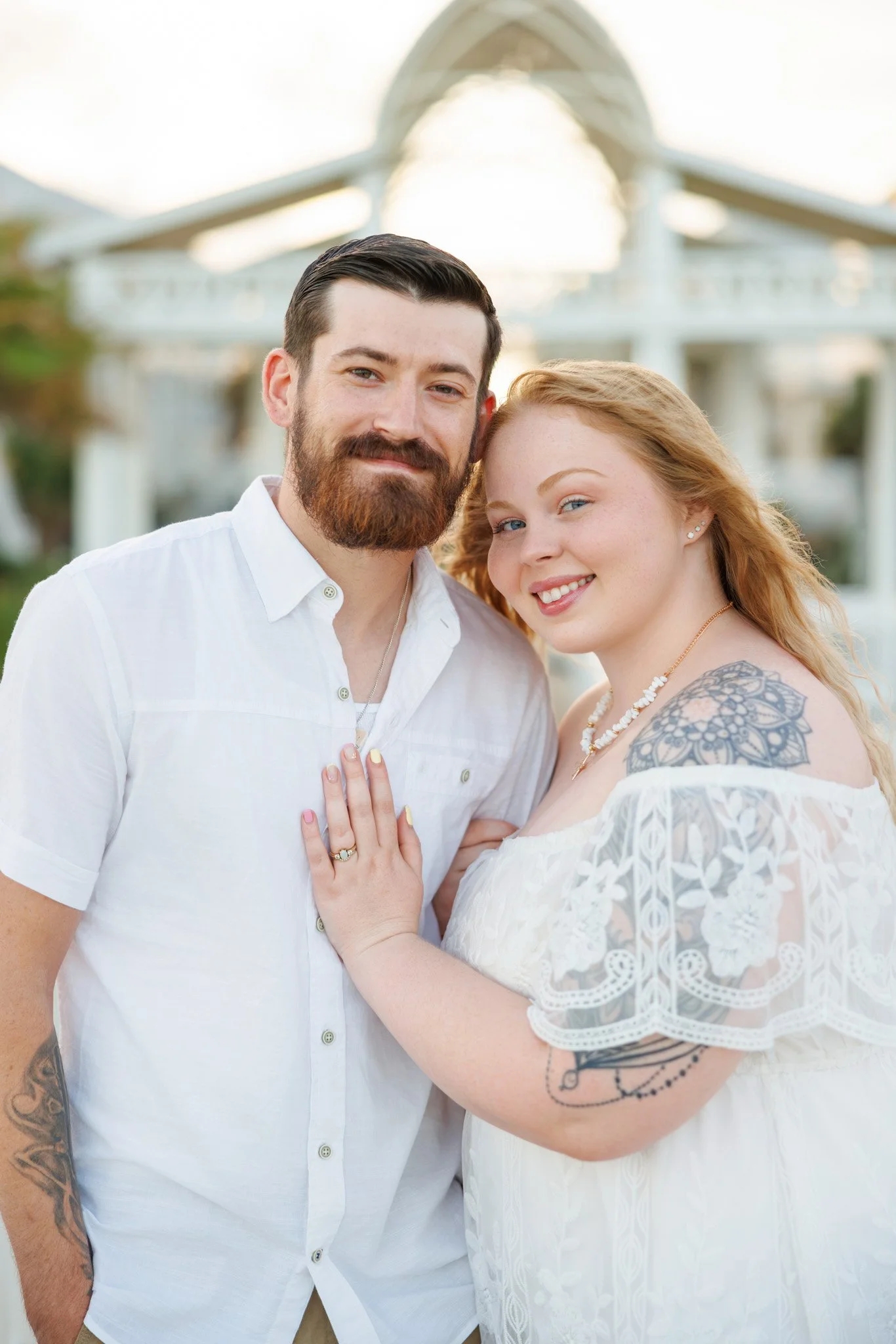 A couple standing outdoors near a large white structure, smiling and looking at the camera, with the woman showing her left hand with rings, and both wearing white clothing.