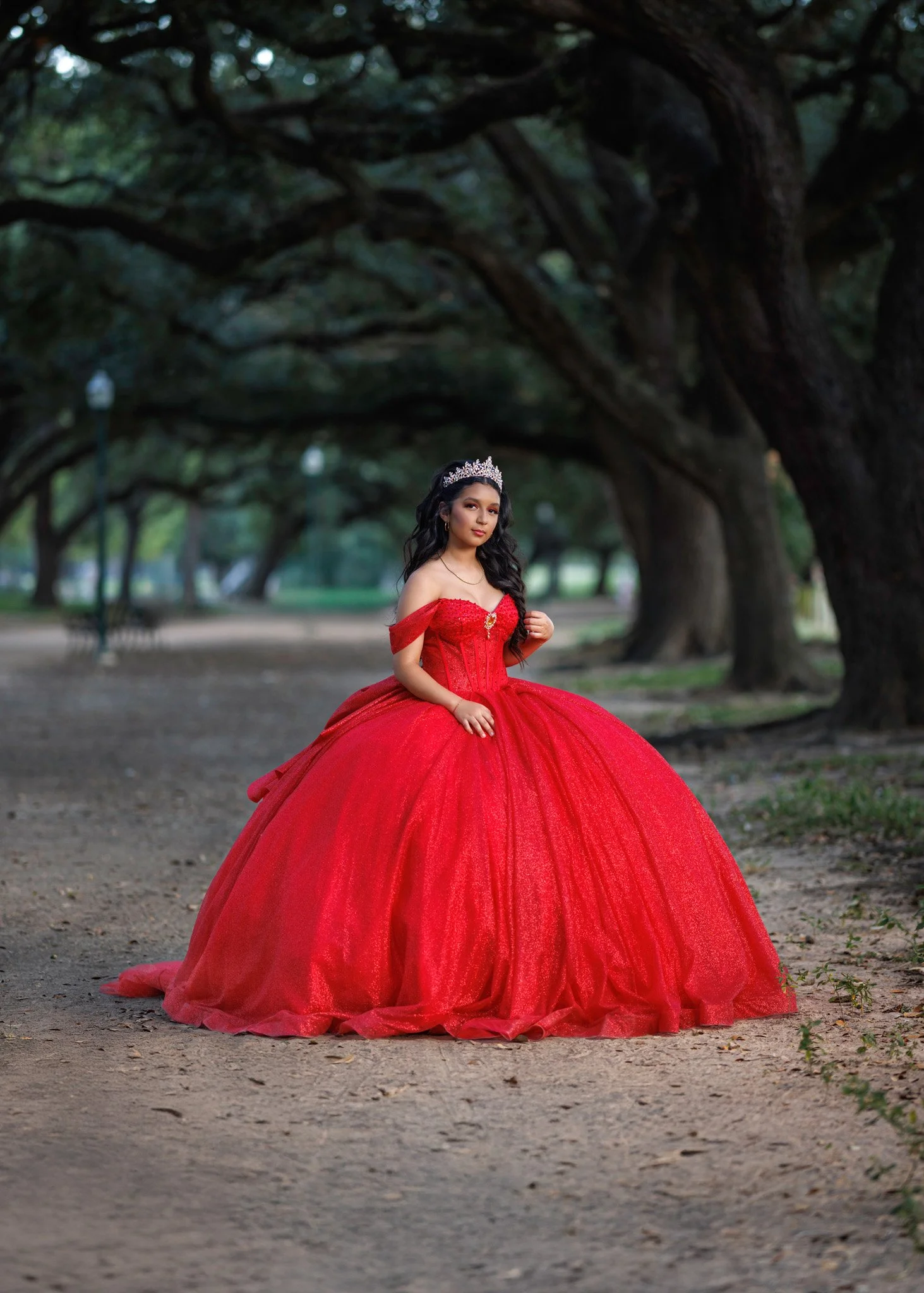 A young woman wearing a red ball gown with off-the-shoulder sleeves, sitting outdoors in a park with large trees and a dirt ground, wearing a tiara and looking at the camera.