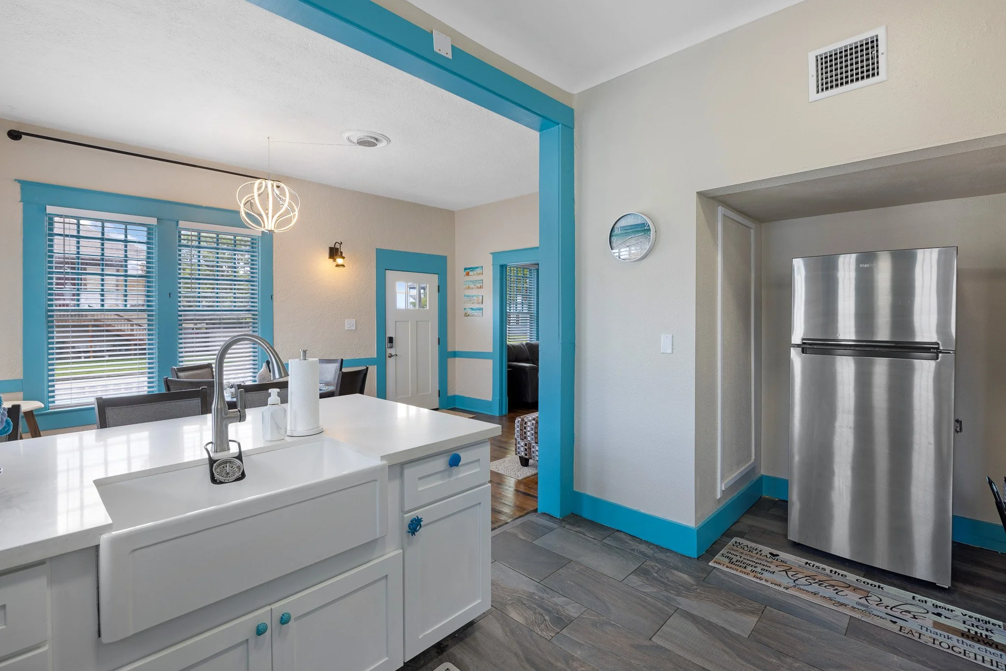 Kitchen with white cabinets, a large white island sink, blue window frames, and a stainless steel fridge. The walls and trim are painted blue, with cream walls and dark wood flooring.