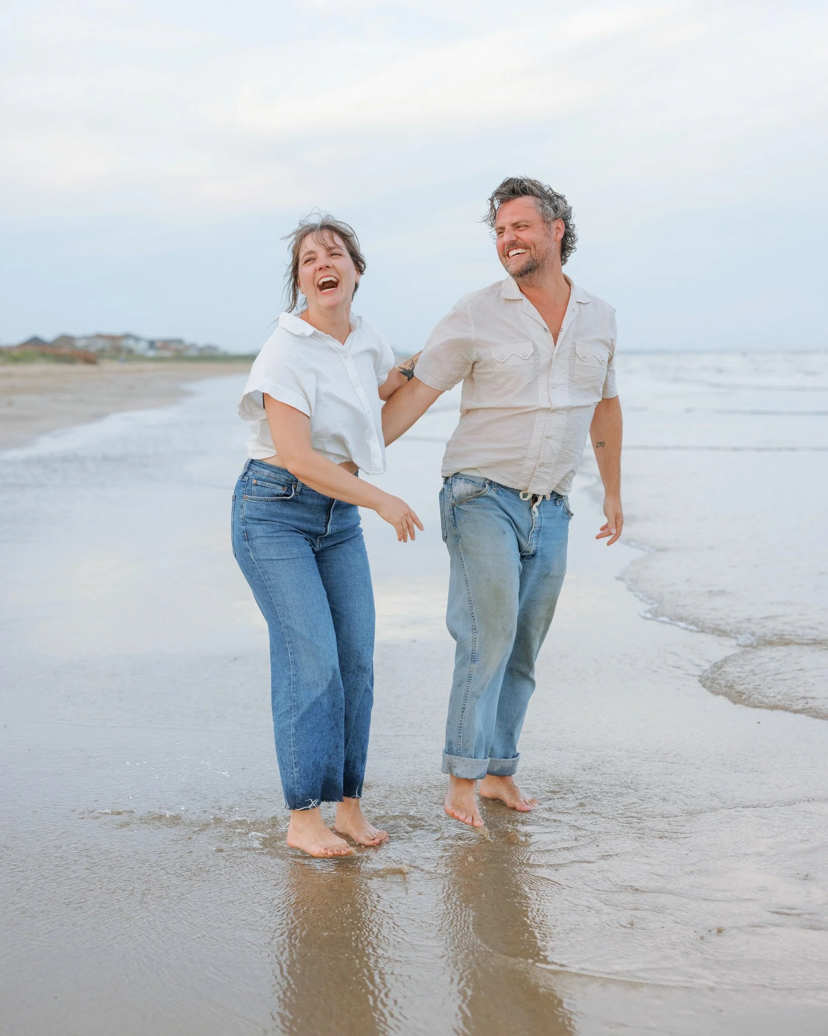 A man and a woman laughing and walking barefoot along the shoreline at the beach.