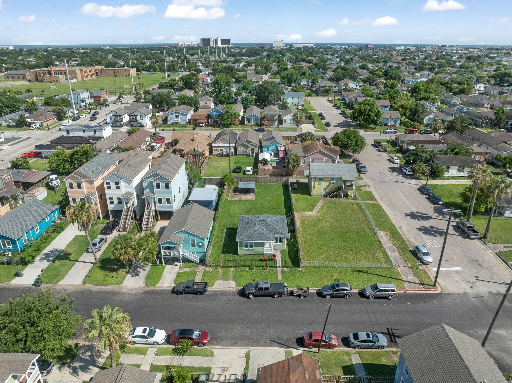Aerial view of a residential neighborhood with houses, streets, parking, and greenery, with a city skyline in the distance.