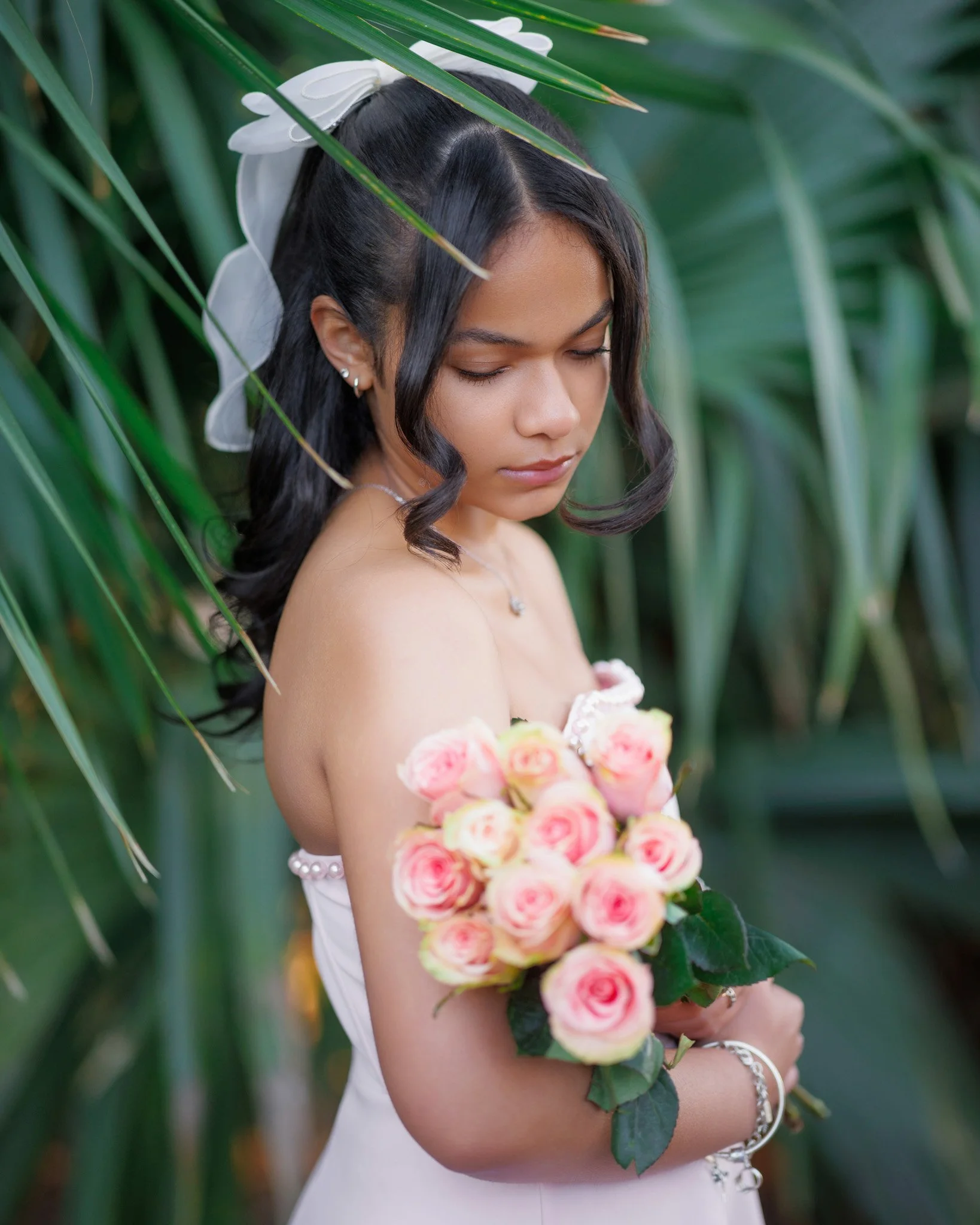 A woman with dark hair styled in loose curls, wearing earrings and a pearl bracelet, holds a bouquet of pink and peach roses while standing among green tropical plants with large leaves.