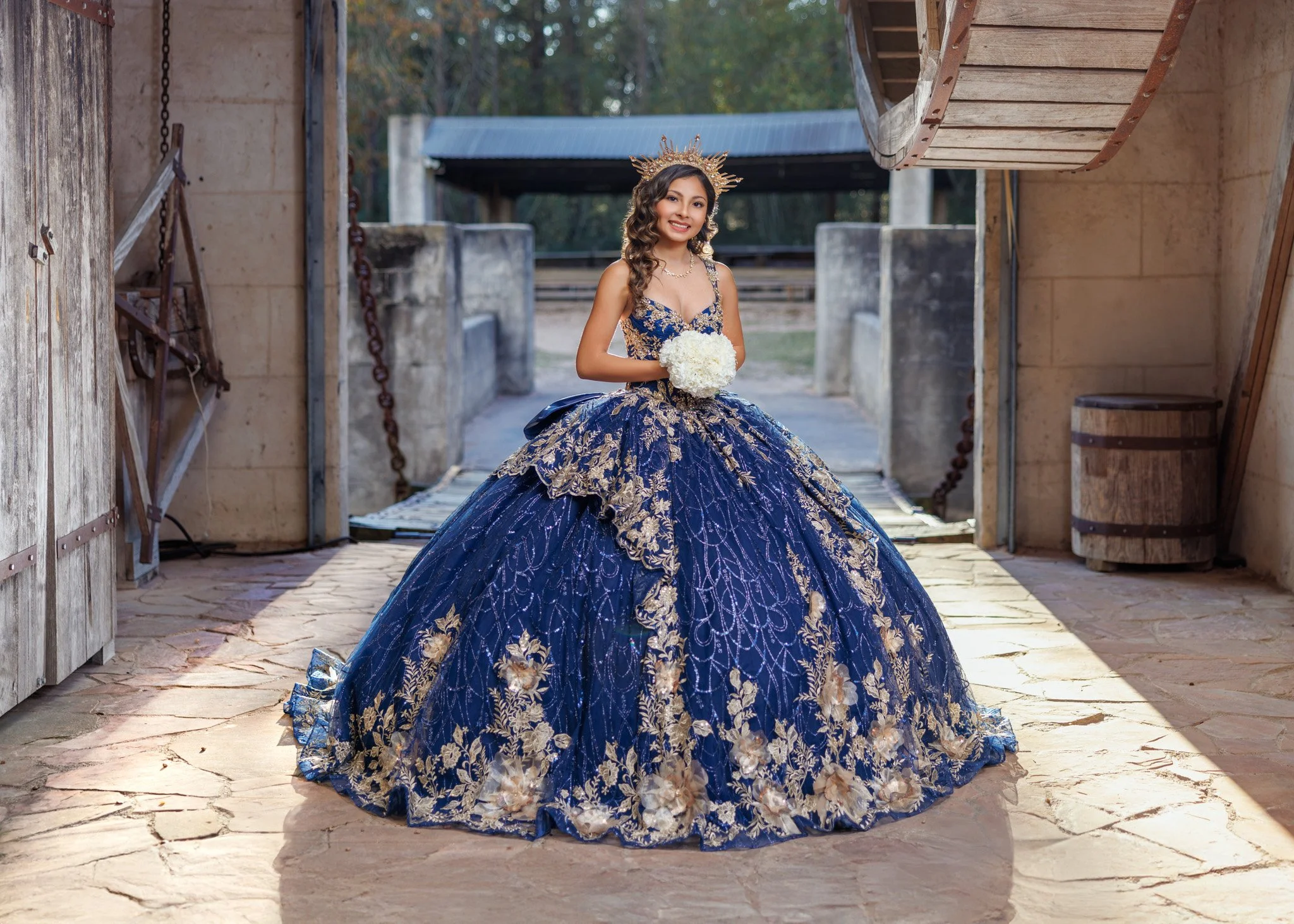 Young woman in an elaborate navy blue ball gown with gold embroidery, holding a bouquet of white flowers, standing in an outdoor setting with stone pavement, wooden structures, and trees in the background.