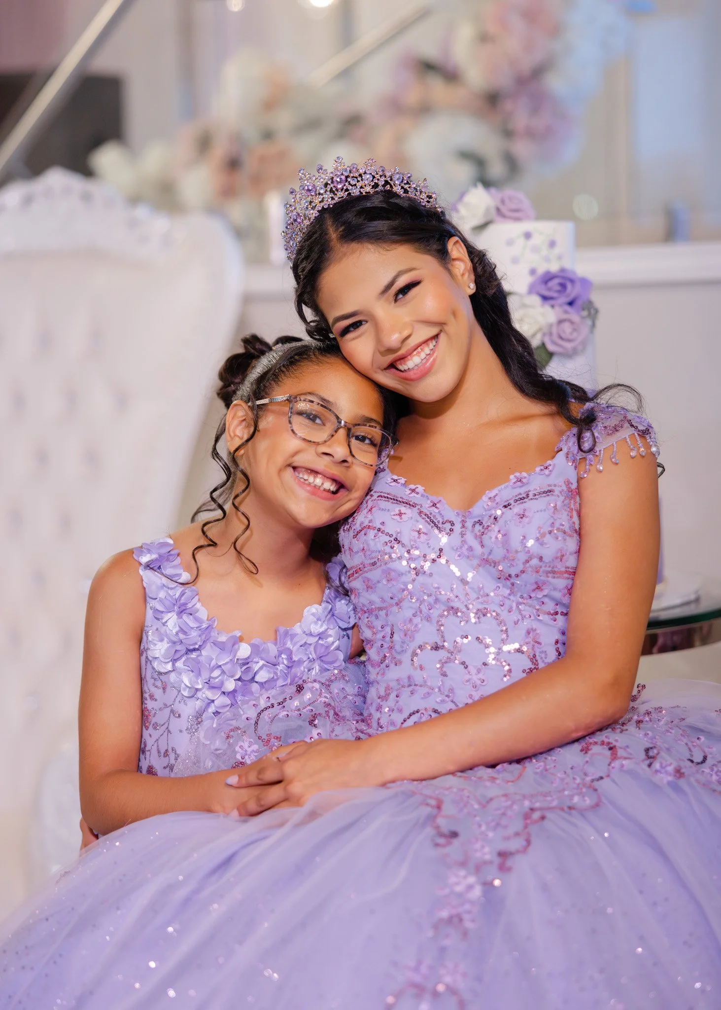 A young girl and a woman, possibly a mother and daughter, dressed in matching lavender dresses with floral accents, sitting together at a celebration. The woman is wearing a tiara and both are smiling happily.