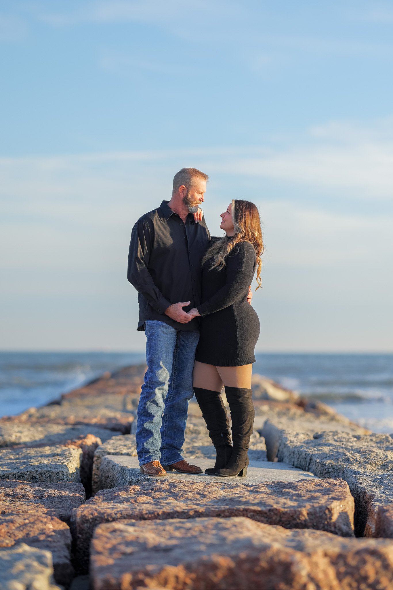 A couple standing close together on rocks by the ocean, looking into each other's eyes during sunset.
