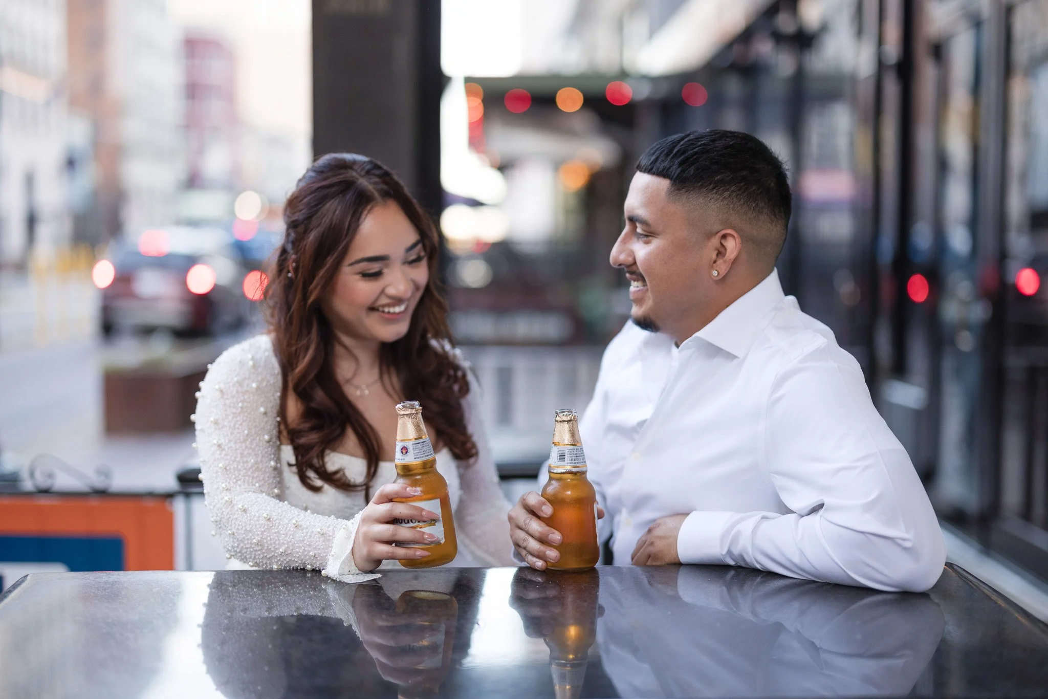A man and woman smiling and holding beer bottles, sitting at a reflective table outdoors in an urban setting, with city buildings and blurred traffic lights in the background.
