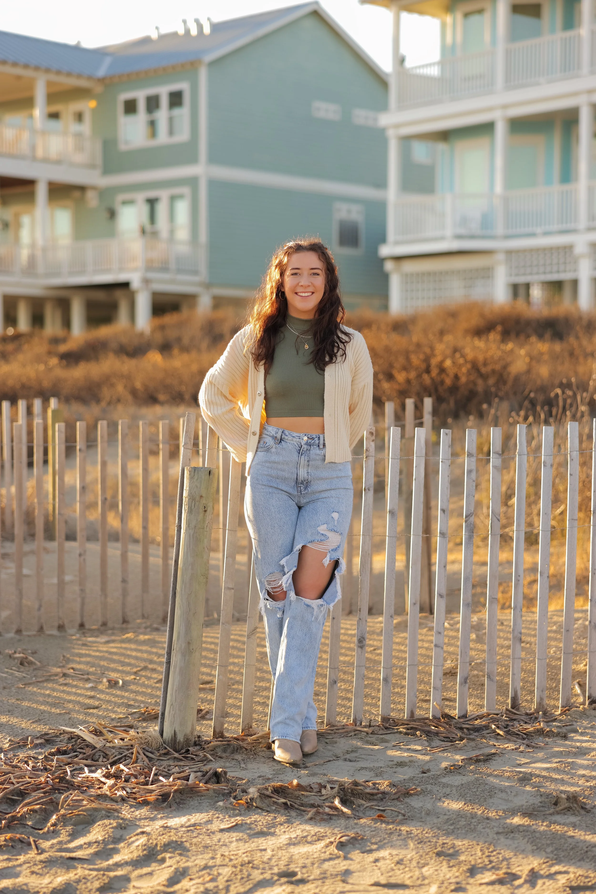 Young woman with curly hair smiling, standing on a sandy beach in front of a wooden fence, with two-story residential houses in the background during sunset.