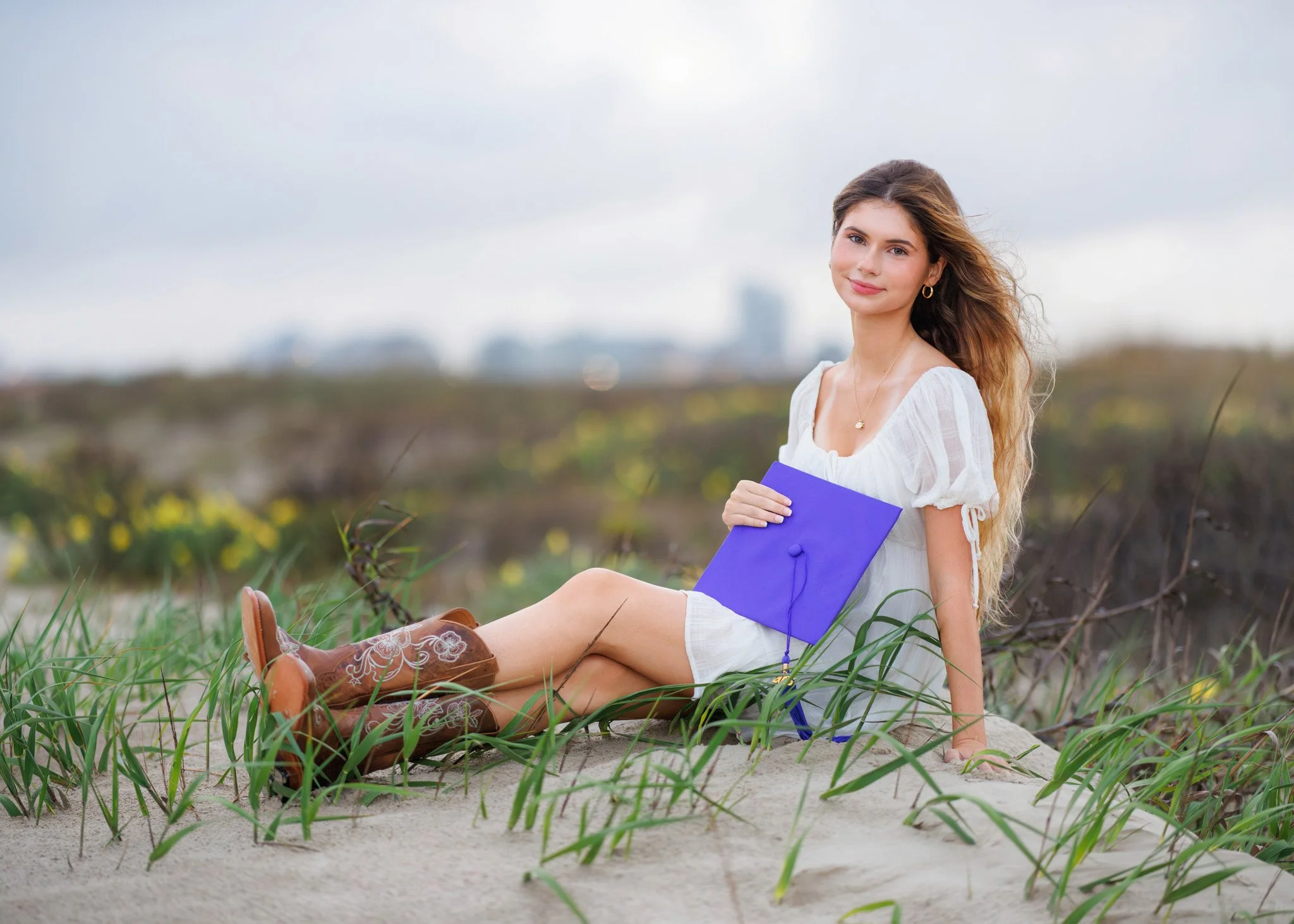 Young woman sitting on sandy beach with green grass, holding a purple diploma cover, wearing a white dress and brown boots, with a cityscape in the background.