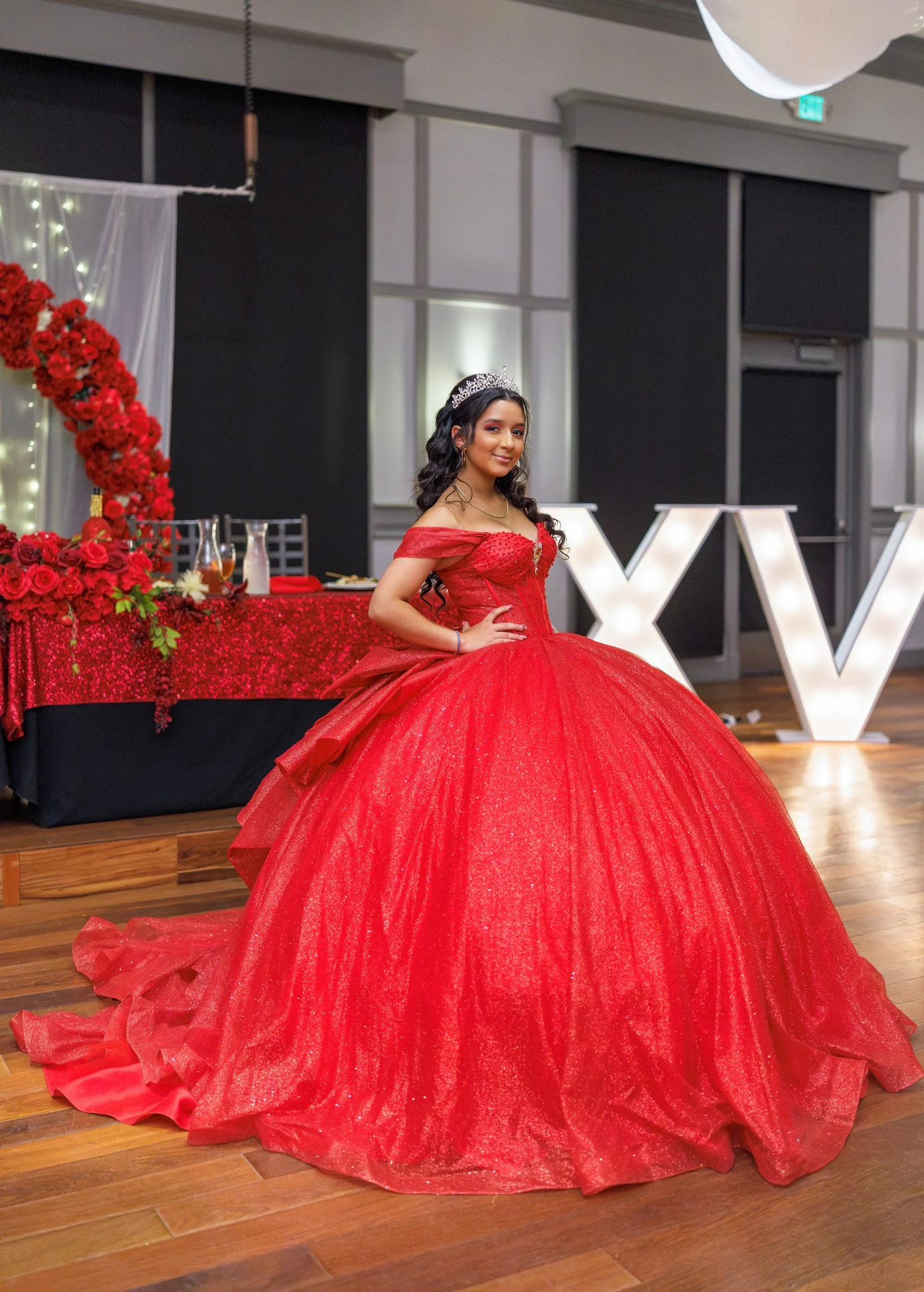 A young woman wearing a red formal gown and a tiara, standing at a decorated event venue with a table decorated with red roses and a large illuminated V in the background.