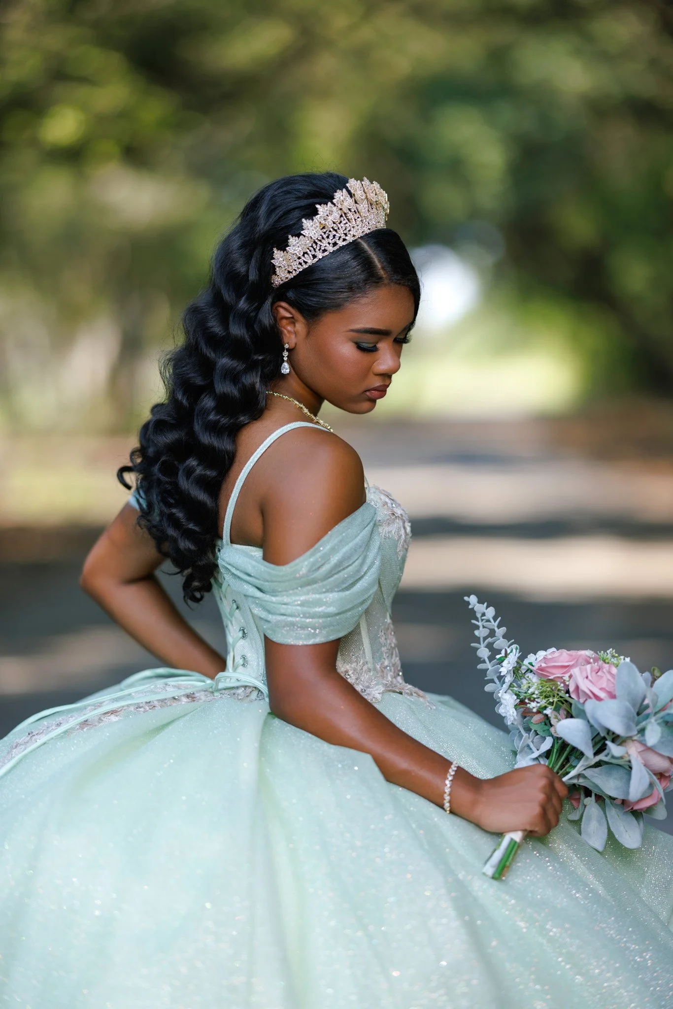 Young woman dressed as a princess in a sparkling light green gown, wearing a tiara, holding a bouquet of pink flowers, outdoors with blurred green trees in background.