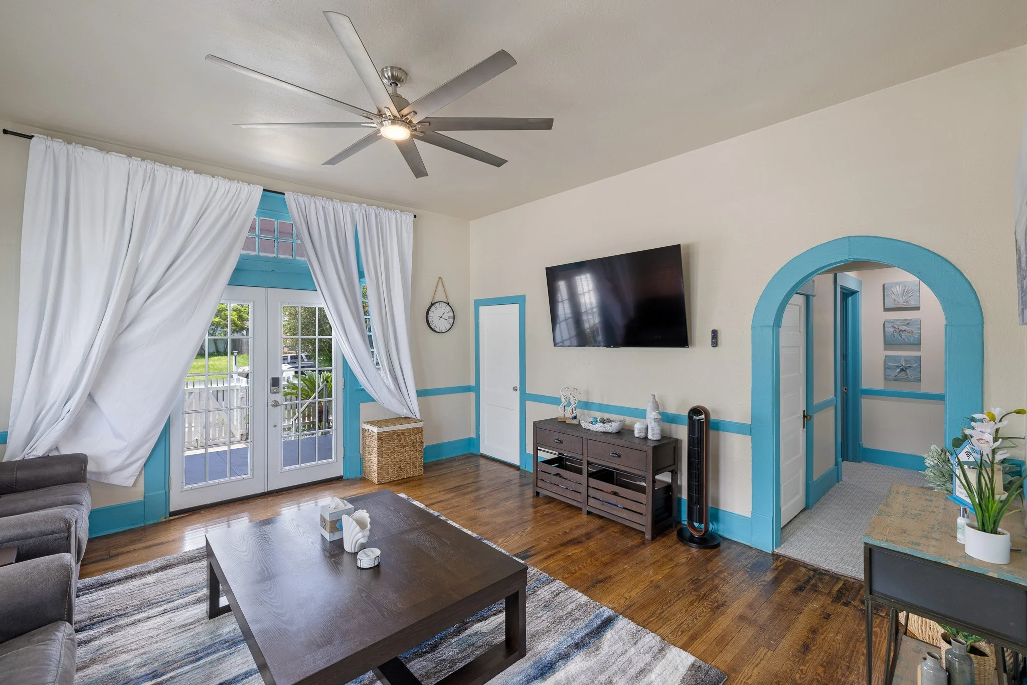 Living room with large glass door and white curtains, wooden floor, wall-mounted TV, gray sofa, coffee table, blue trim accents, wicker basket, and decorative items.