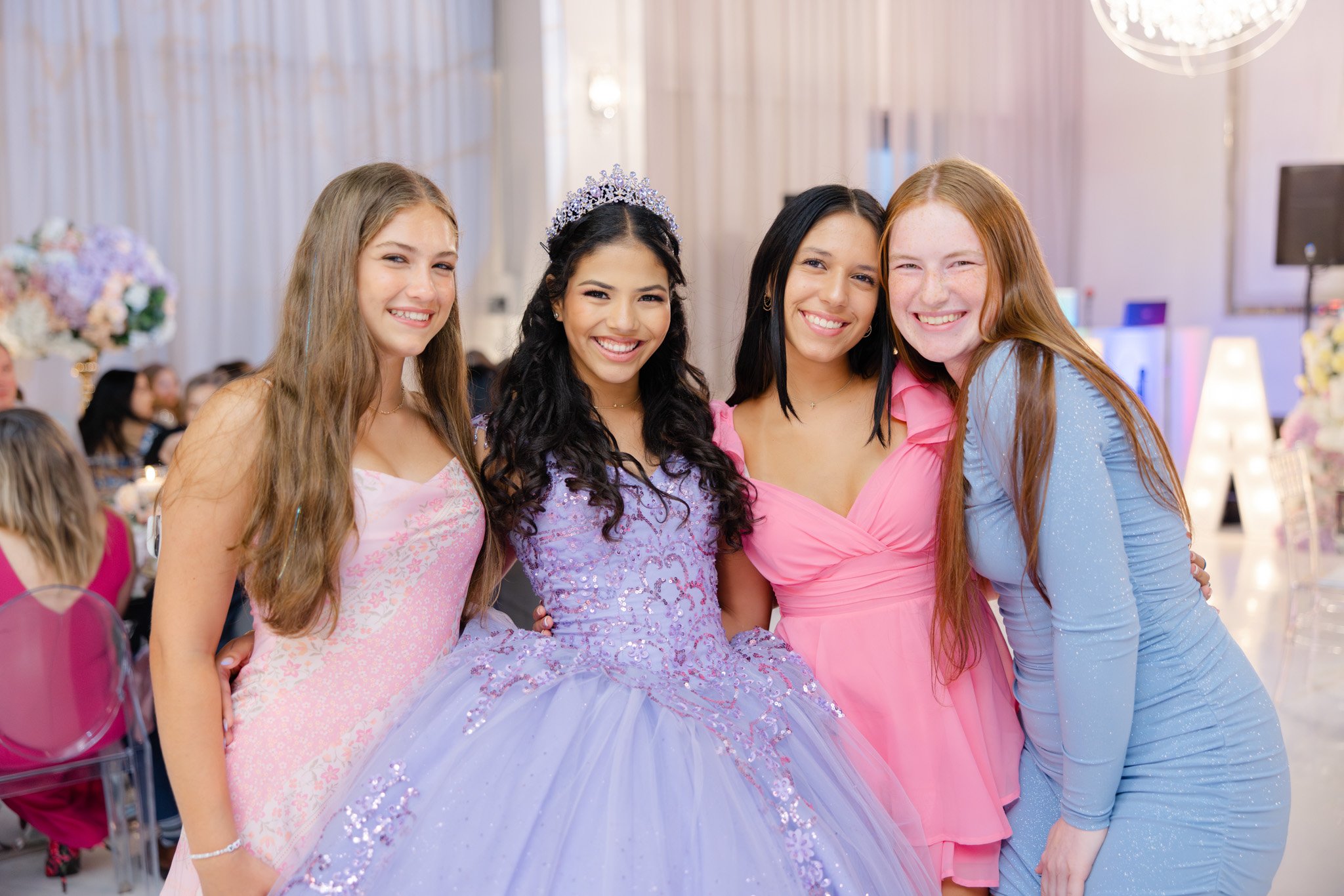Four young women smiling at a quinceañera celebration, with one wearing a princess-like lavender dress and tiara, another in a pink dress, another in a pink dress, and the last in a blue dress.
