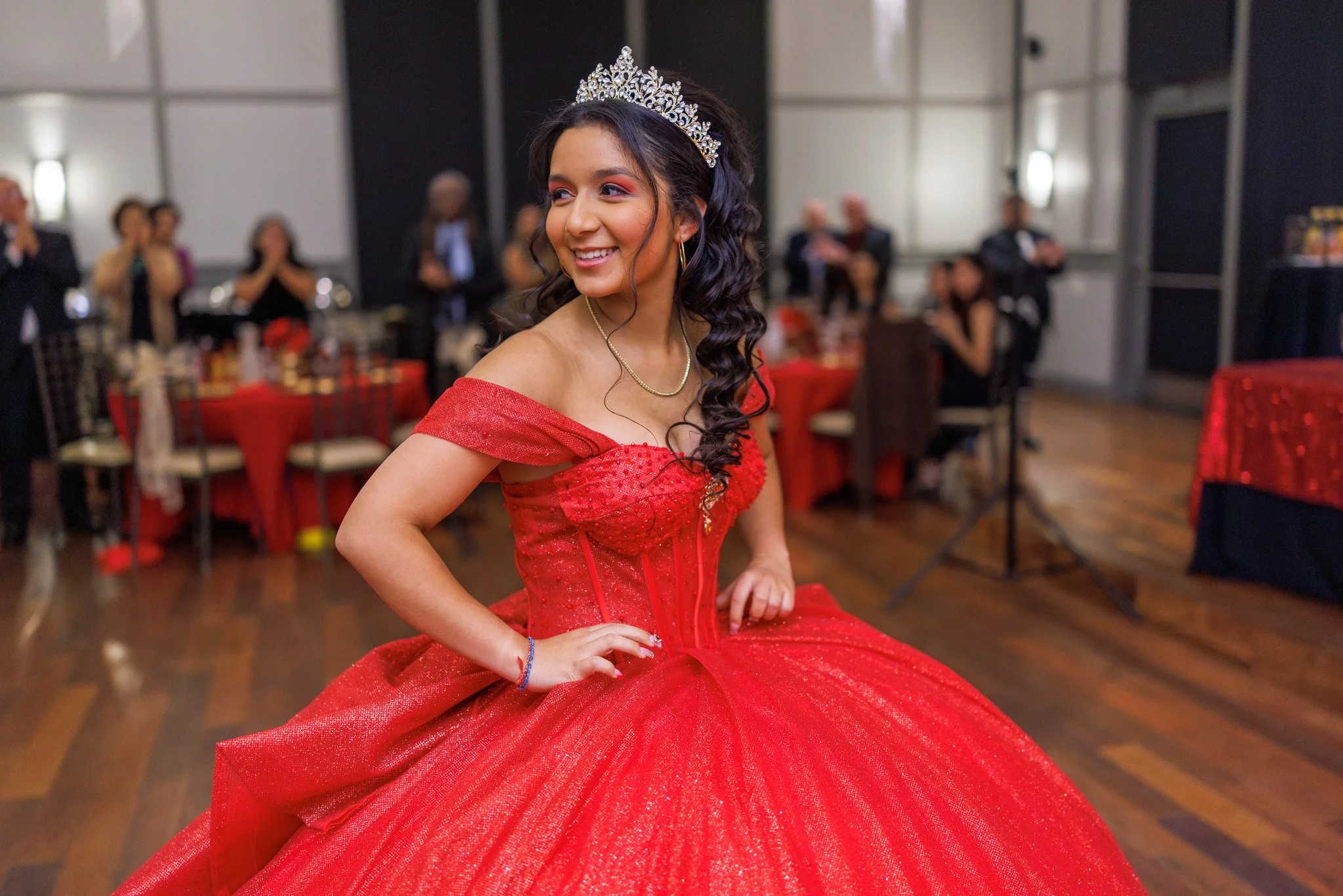 A young woman in a red formal gown and a tiara, smiling at a celebration or party event.