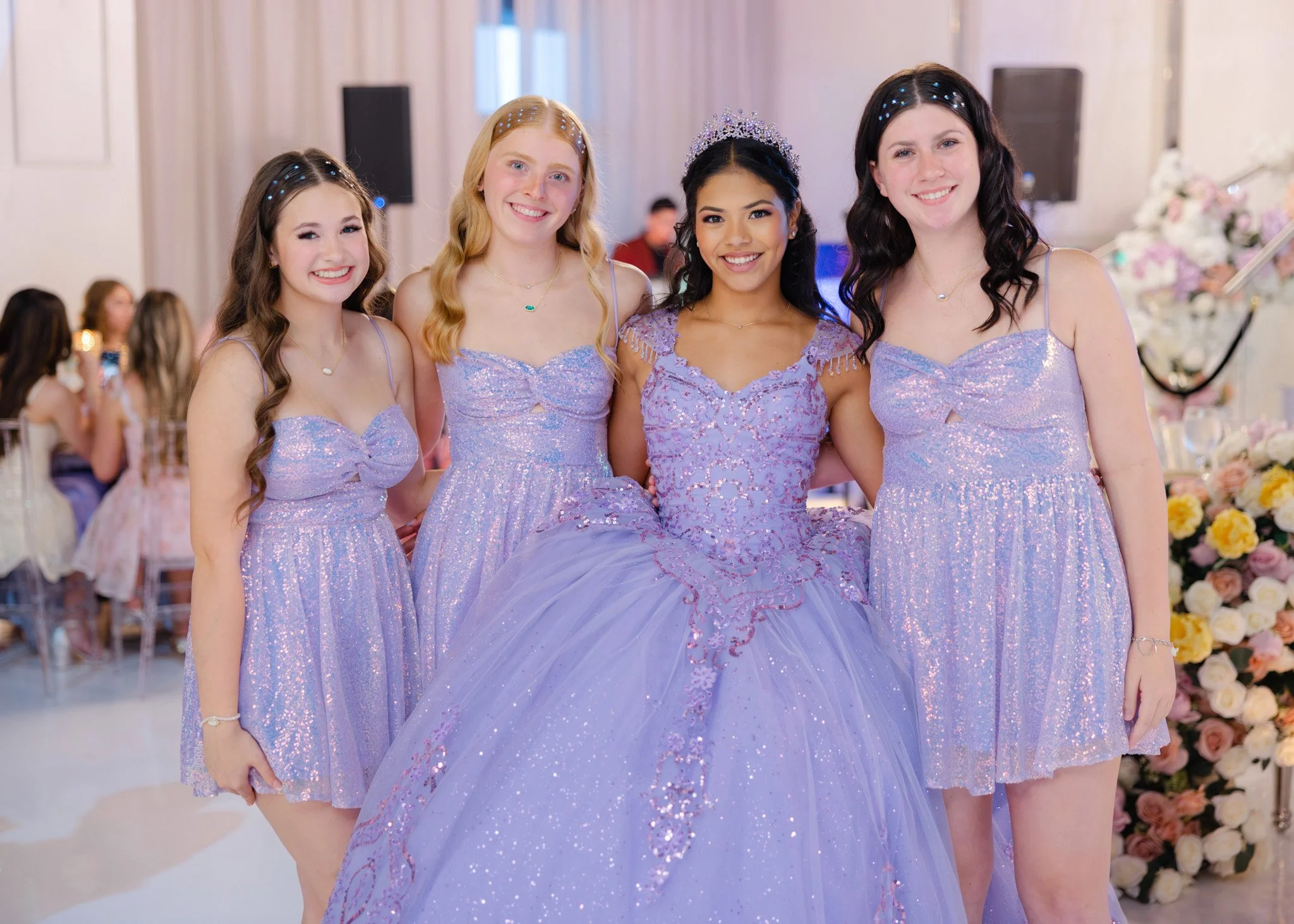 Four young women dressed in lavender dresses standing together at an indoor event, smiling at the camera.
