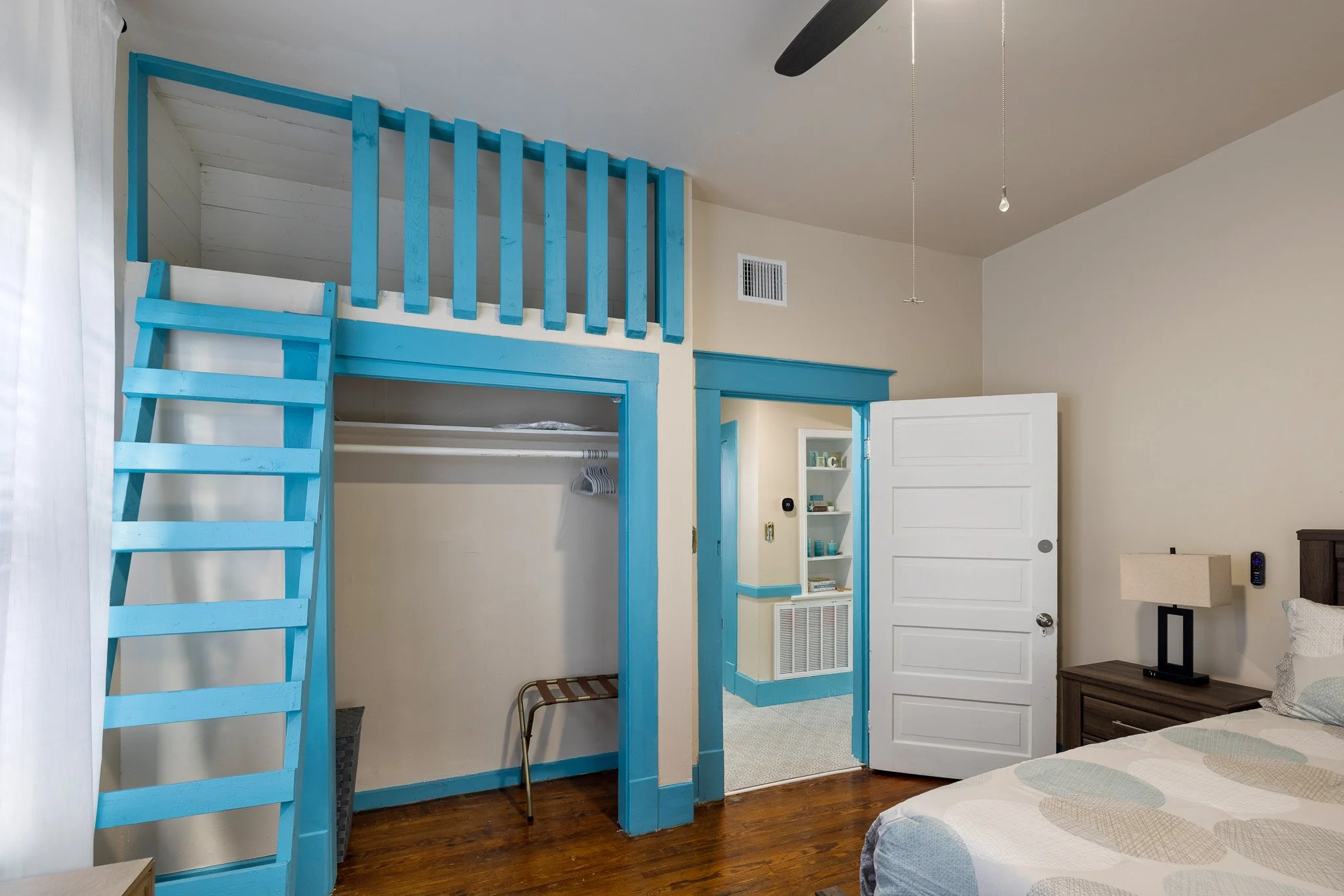 Interior of a bedroom with blue and white accents, a loft area with a ladder, an open closet, a white door, a nightstand with a lamp, and a bed with a patterned bedspread.