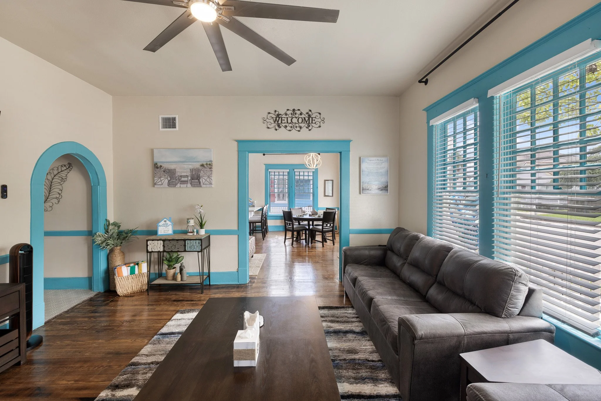Living room with a gray couch, wooden coffee table, large windows with blinds, blue trim, and a view into a dining area with a round table and four chairs.