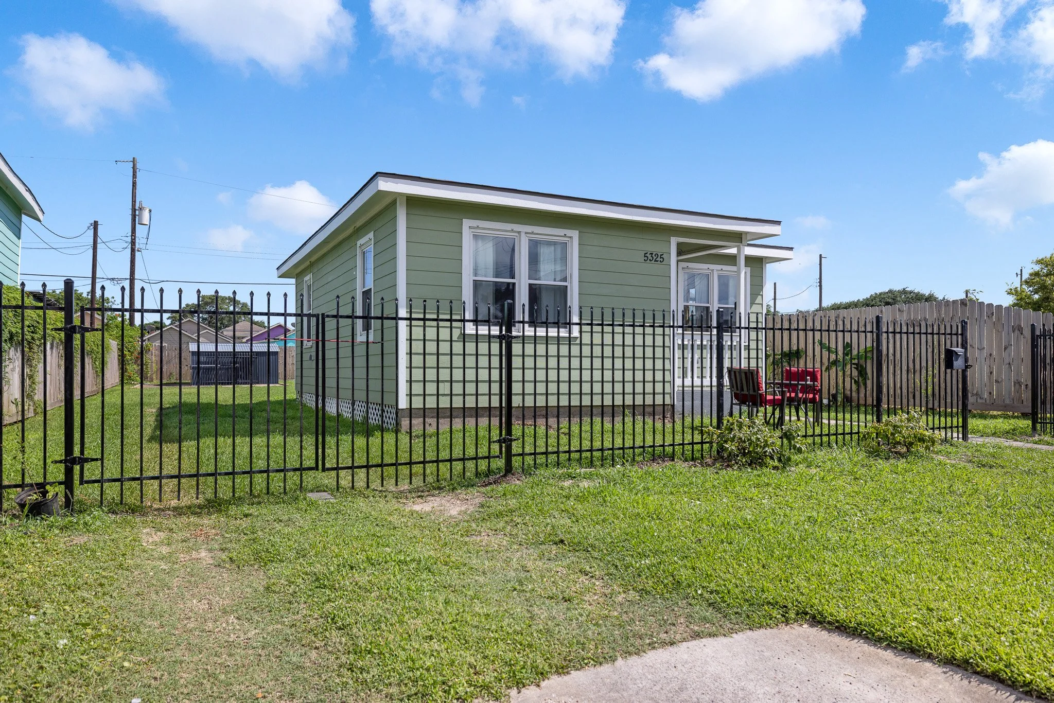 A small, light green house with white trim and a black metal fence around the yard, featuring a patio with red outdoor furniture.