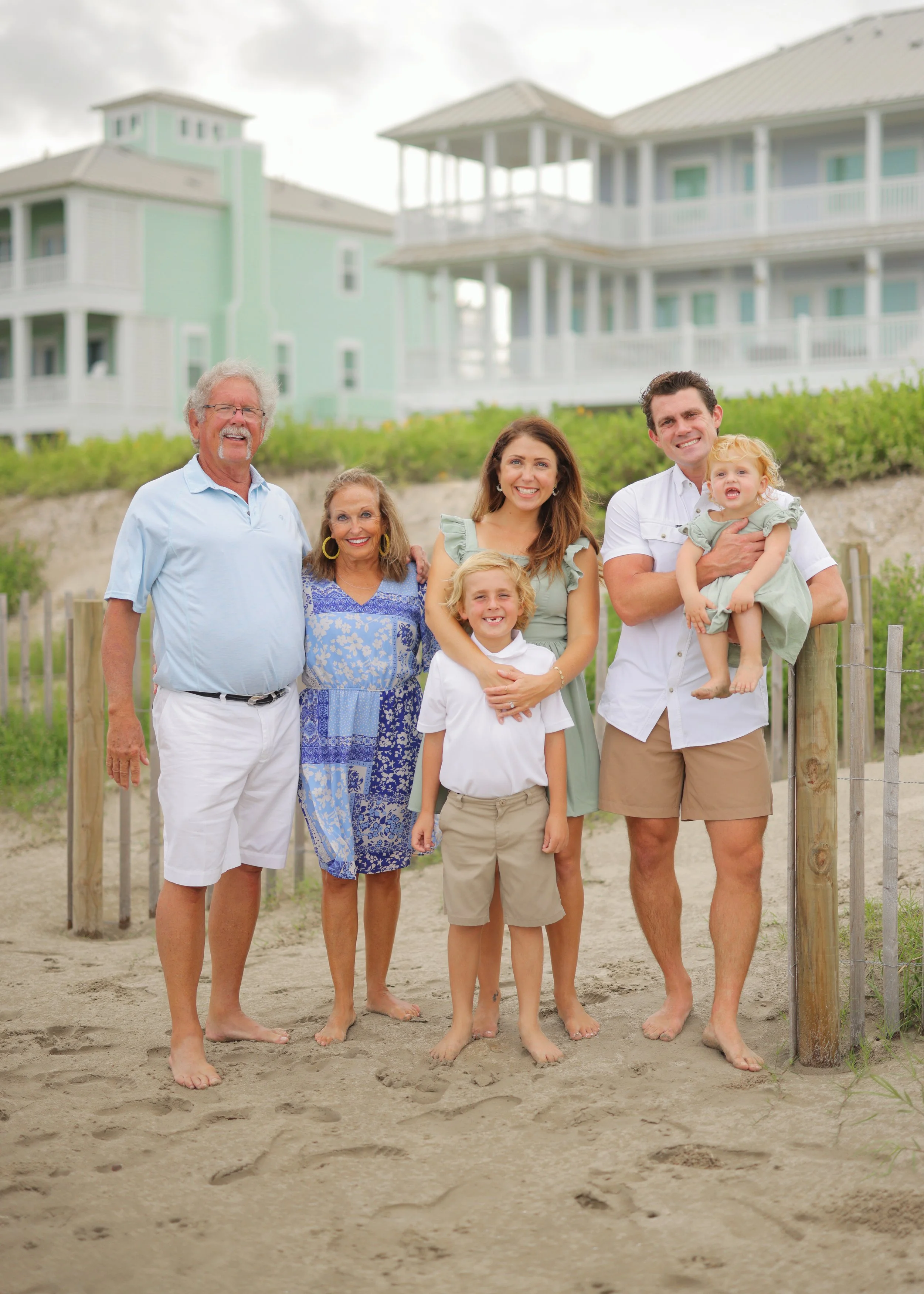 A multi-generational family of six standing barefoot on a sandy beach, smiling, with a background of beach houses and a cloudy sky.