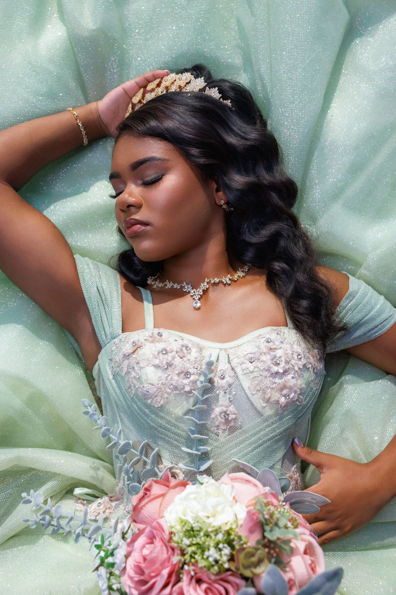 A woman with dark hair styled in waves, wearing a pearl and floral necklace, is lying on a shimmering, light green fabric background, holding a bouquet of pink, white, and purple flowers with greenery.