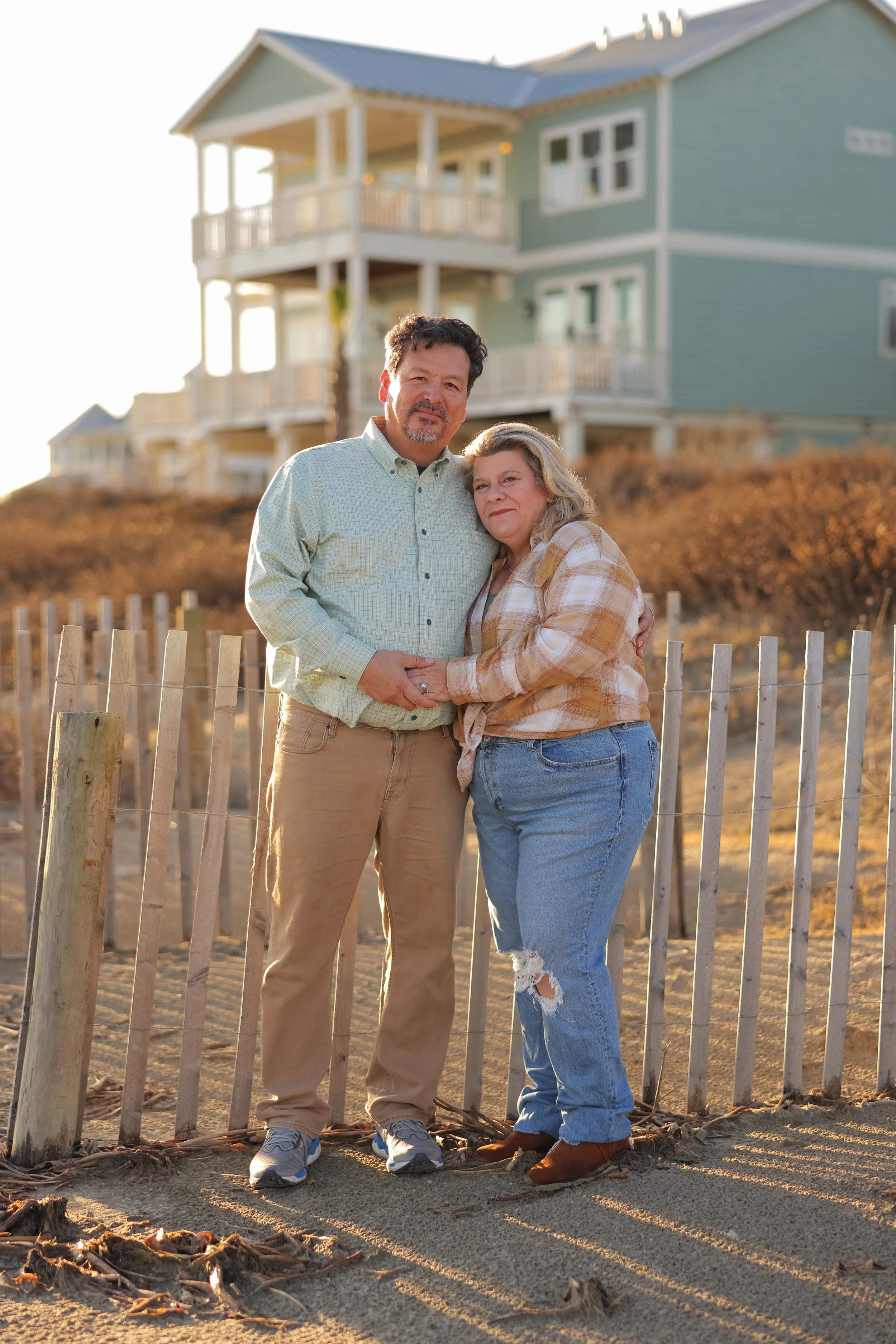 A middle-aged couple stands close together outdoors, holding hands and posing for a photo on a beach at sunset. Behind them are two-story beach houses on stilts, and a wooden fence lines the sandy ground. The man is wearing a light green button-up sh