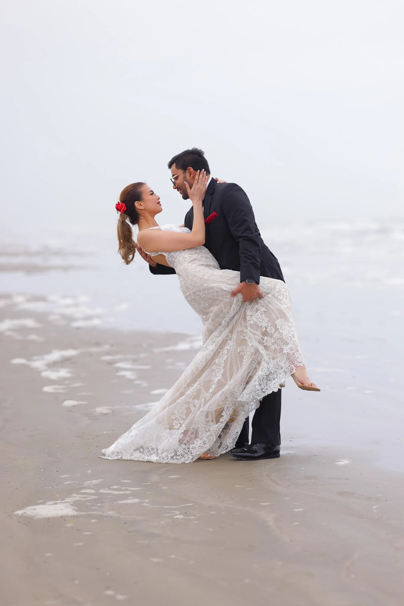 A couple on the beach, with the man in a black tuxedo and the woman in a white wedding dress. The man is holding the woman in his arms, and they are looking into each other's eyes with a scenic ocean background.