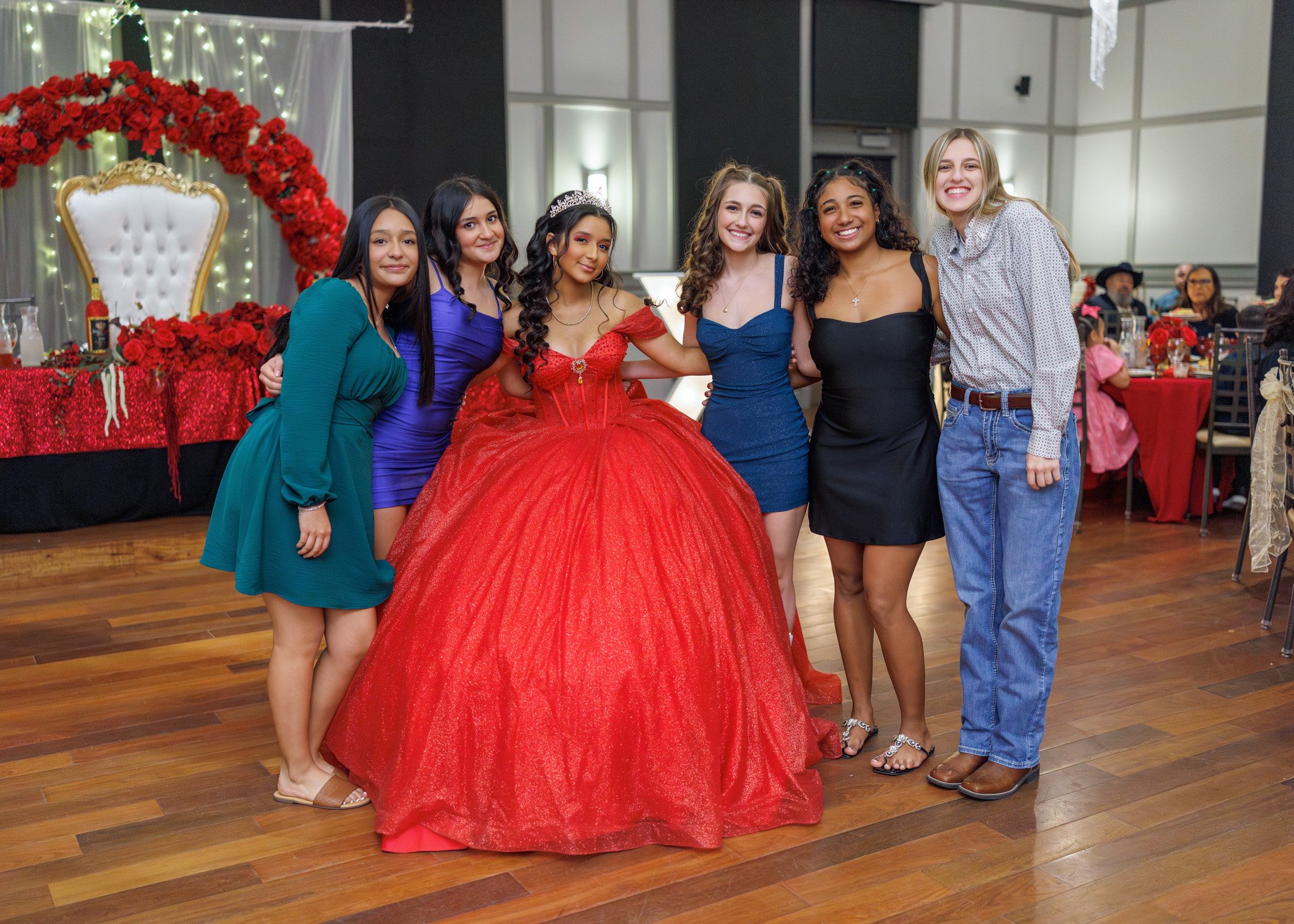 Group of six smiling young women, one wearing a tiara and a red gown, posing together at a celebration or prom in a decorated event hall with a red floral arch and a white and gold chair in the background.