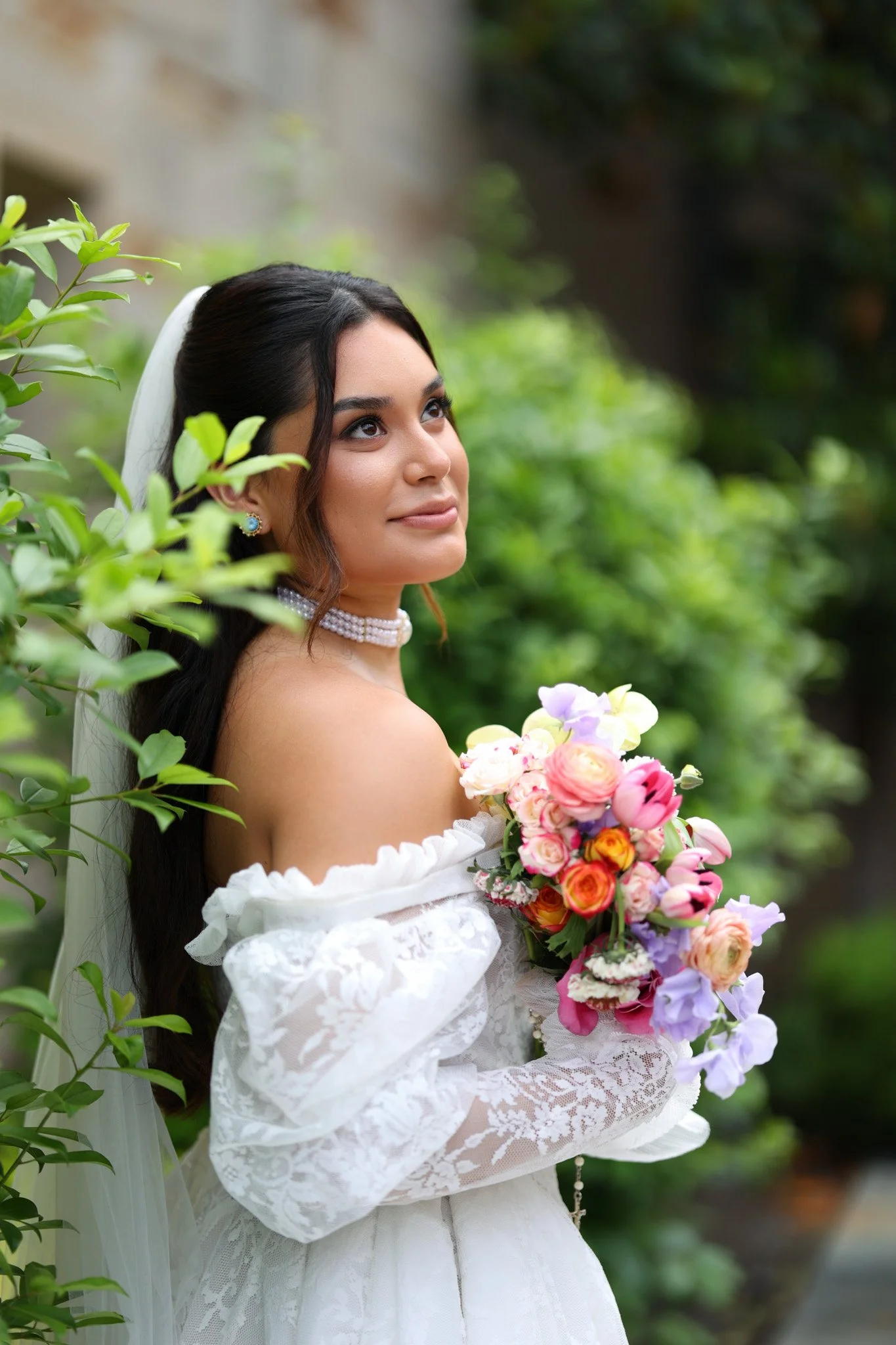 A bride with dark hair and light skin, wearing a white off-shoulder lace wedding dress, holding a colorful bouquet, standing outdoors surrounded by greenery.