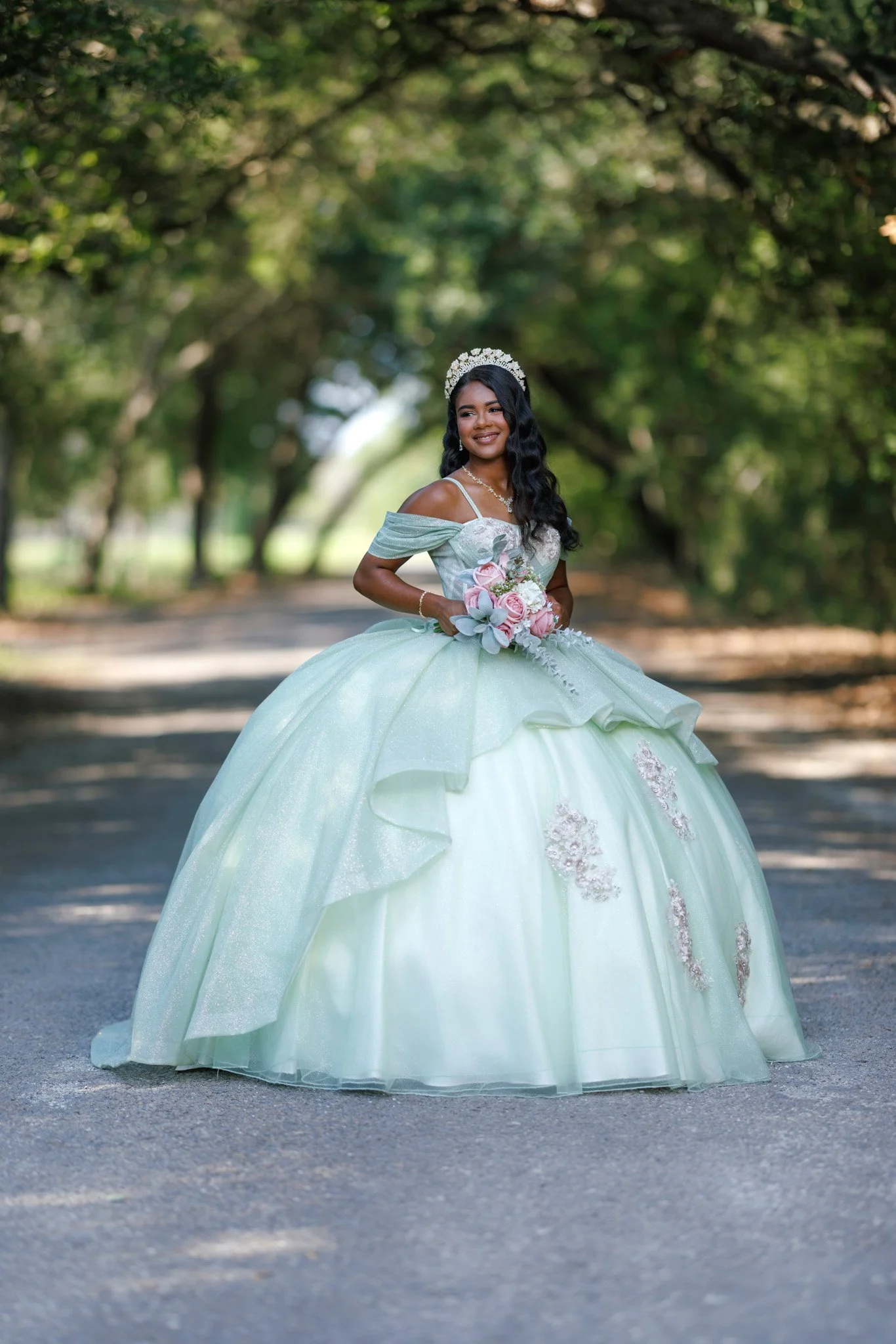 A woman in a light green, ornate ball gown, holding a bouquet of pink and white flowers, standing on a pathway under green arching trees.