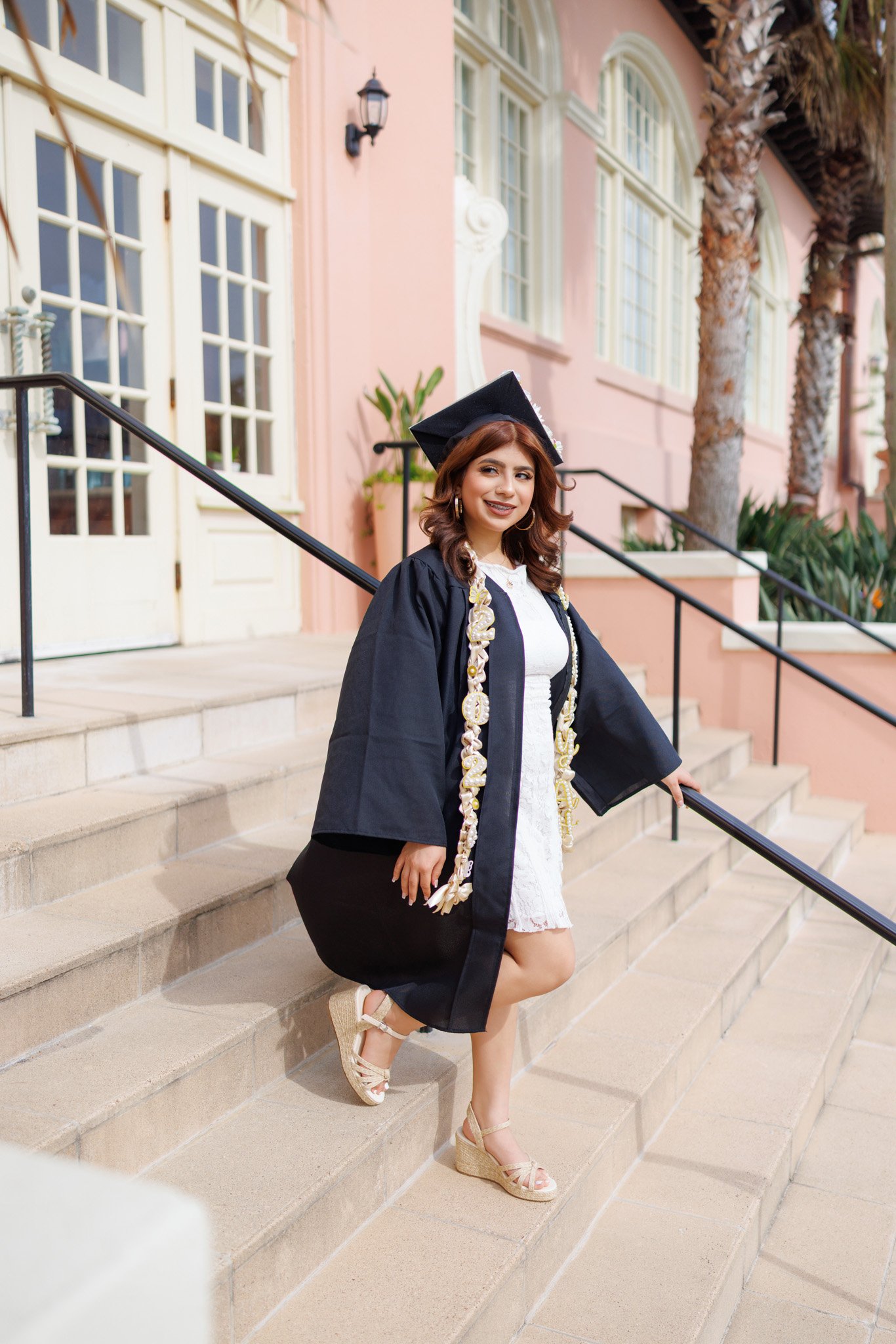 A young woman in a graduation cap and gown stands on stone steps outside a pink building with large windows, palm trees, and greenery in the background.