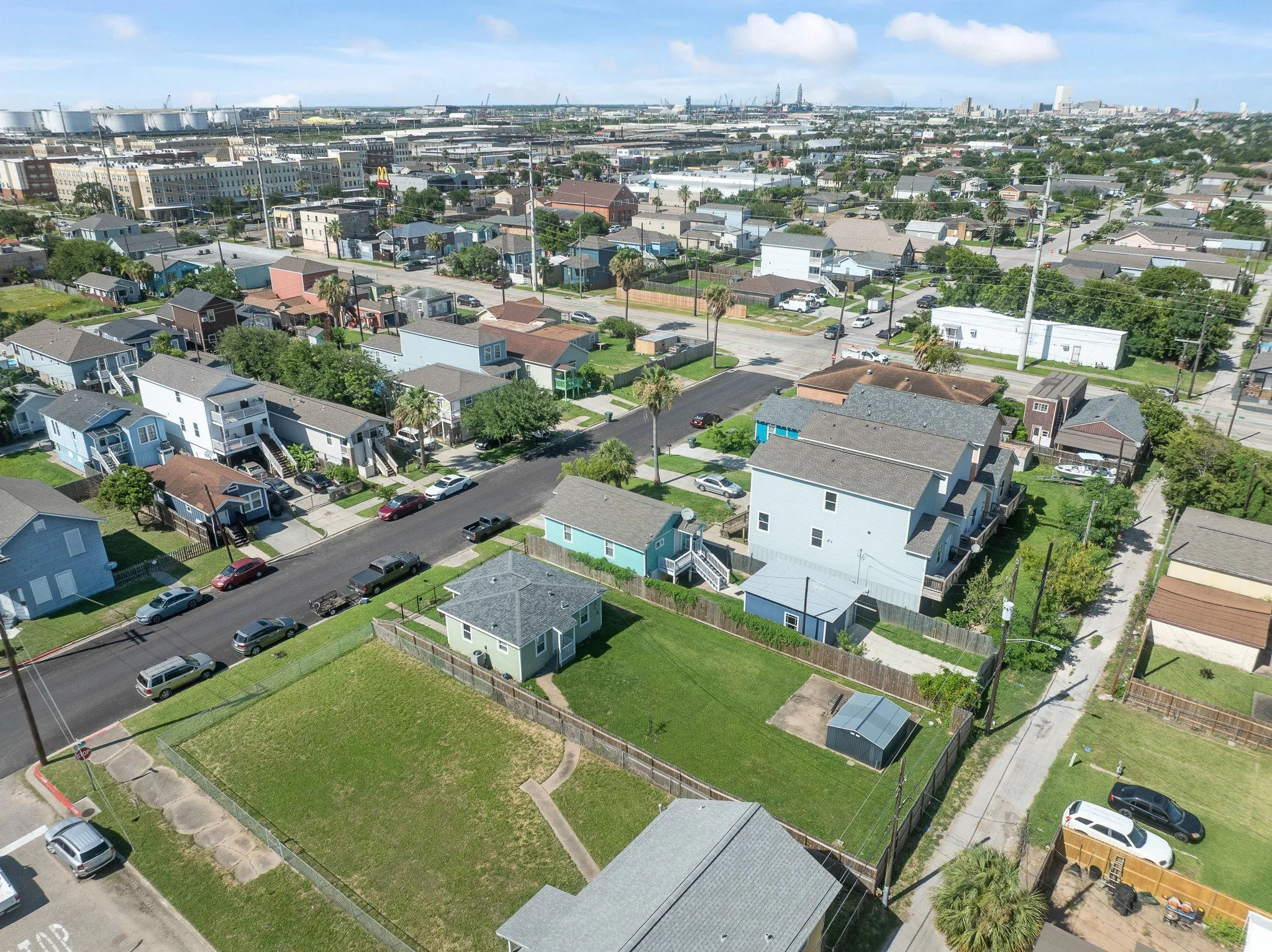 Aerial view of a coastal residential neighborhood with houses, streets, and some cars, with an industrial port and cranes in the background under a partly cloudy sky.