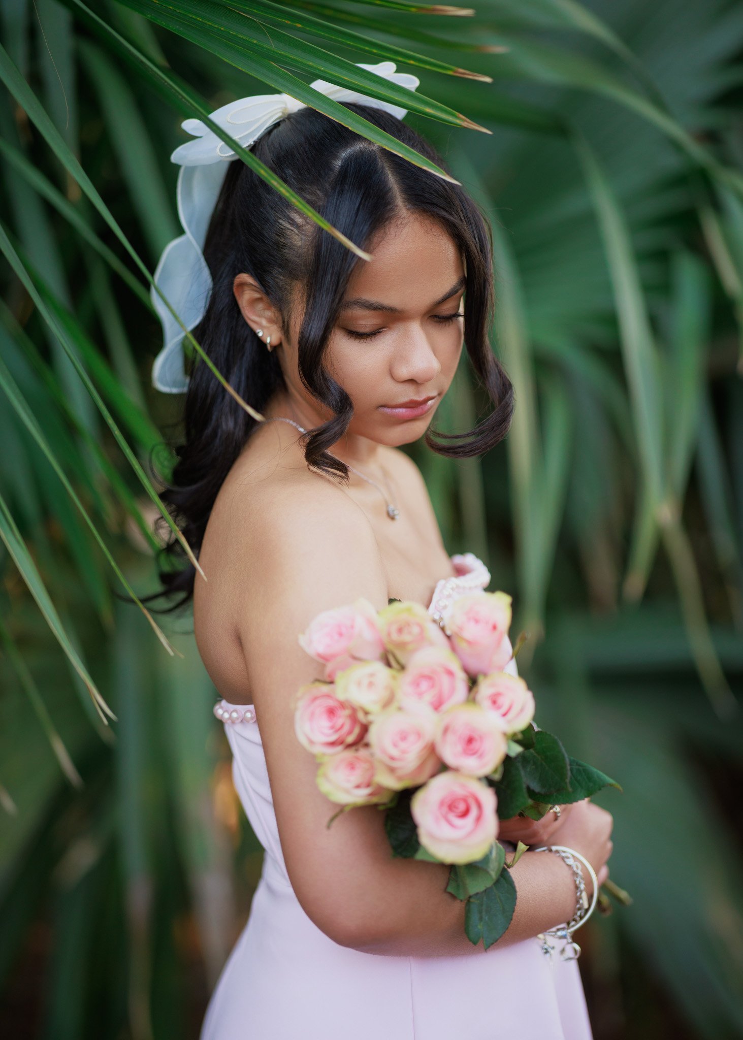 A woman is holding a bouquet of pink roses and wearing a white dress with a floral headpiece. She is surrounded by green tropical plants and has her eyes closed.
