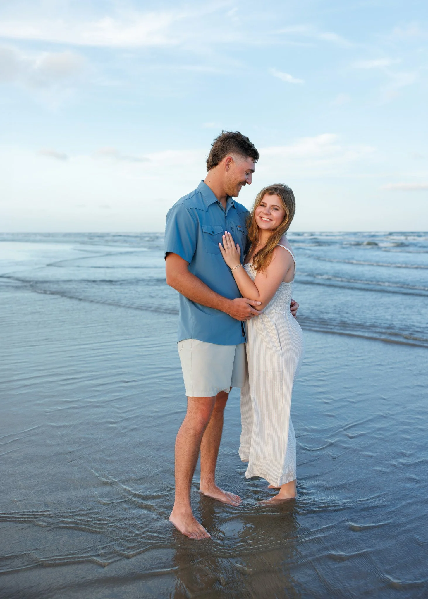 A young couple smiling and embracing while standing in shallow ocean water on a sandy beach, with a cloudy sky in the background.