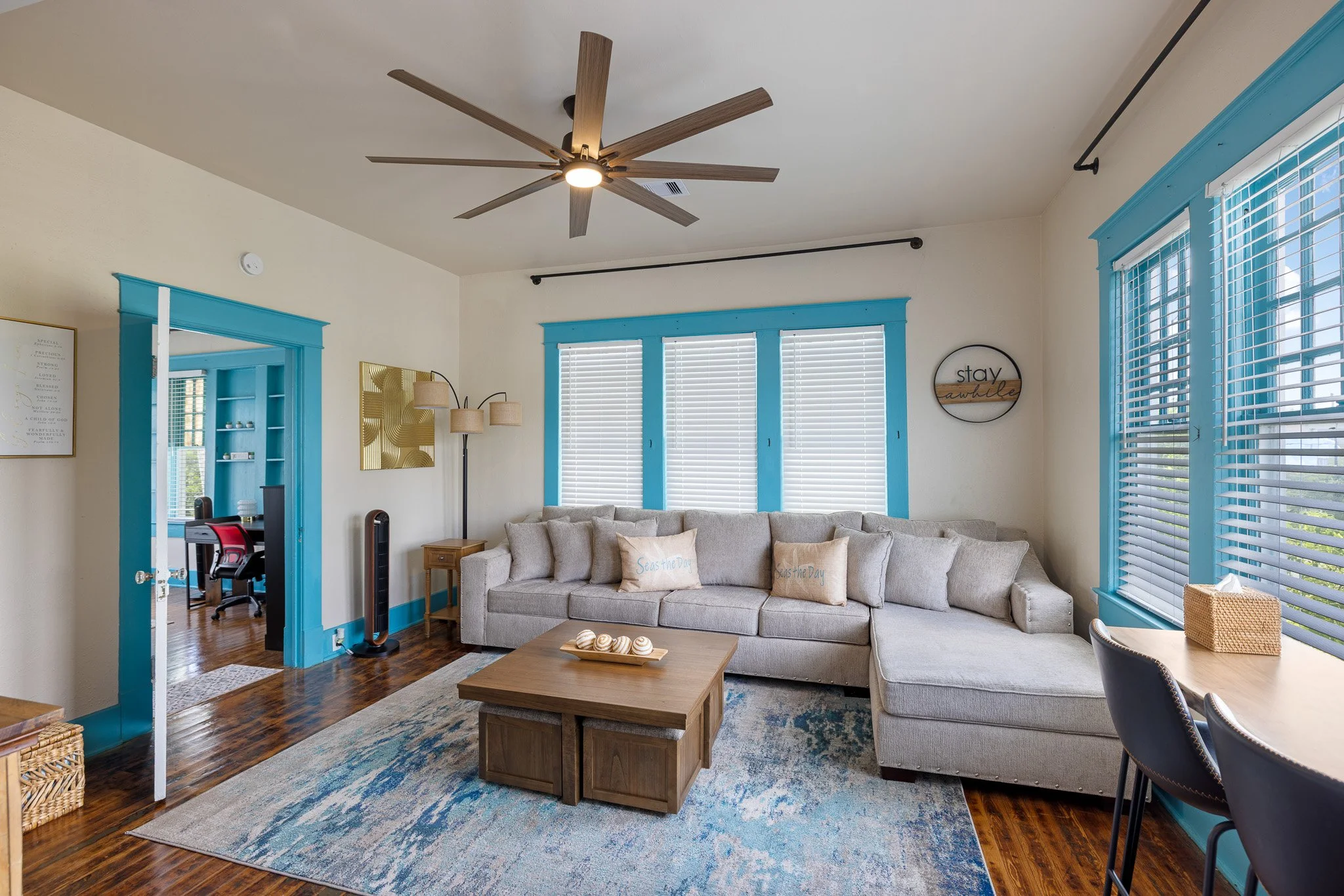Living room with a large beige sectional sofa, a wooden coffee table, and a multi-colored area rug. There are multiple windows with blue trim and white blinds. A ceiling fan with wooden blades is mounted on the ceiling. A light-colored wall has a gol