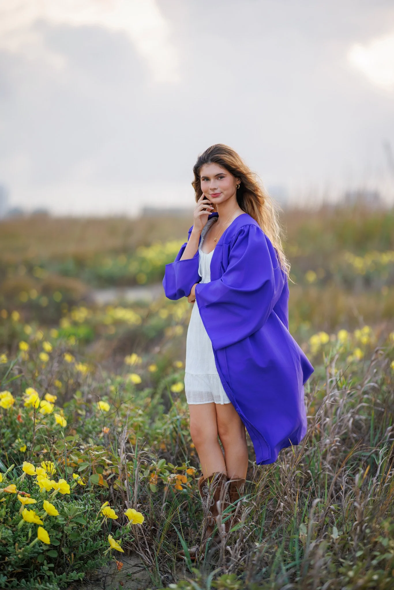 Young woman in a blue dress and jacket standing in a field with yellow flowers, under a cloudy sky.