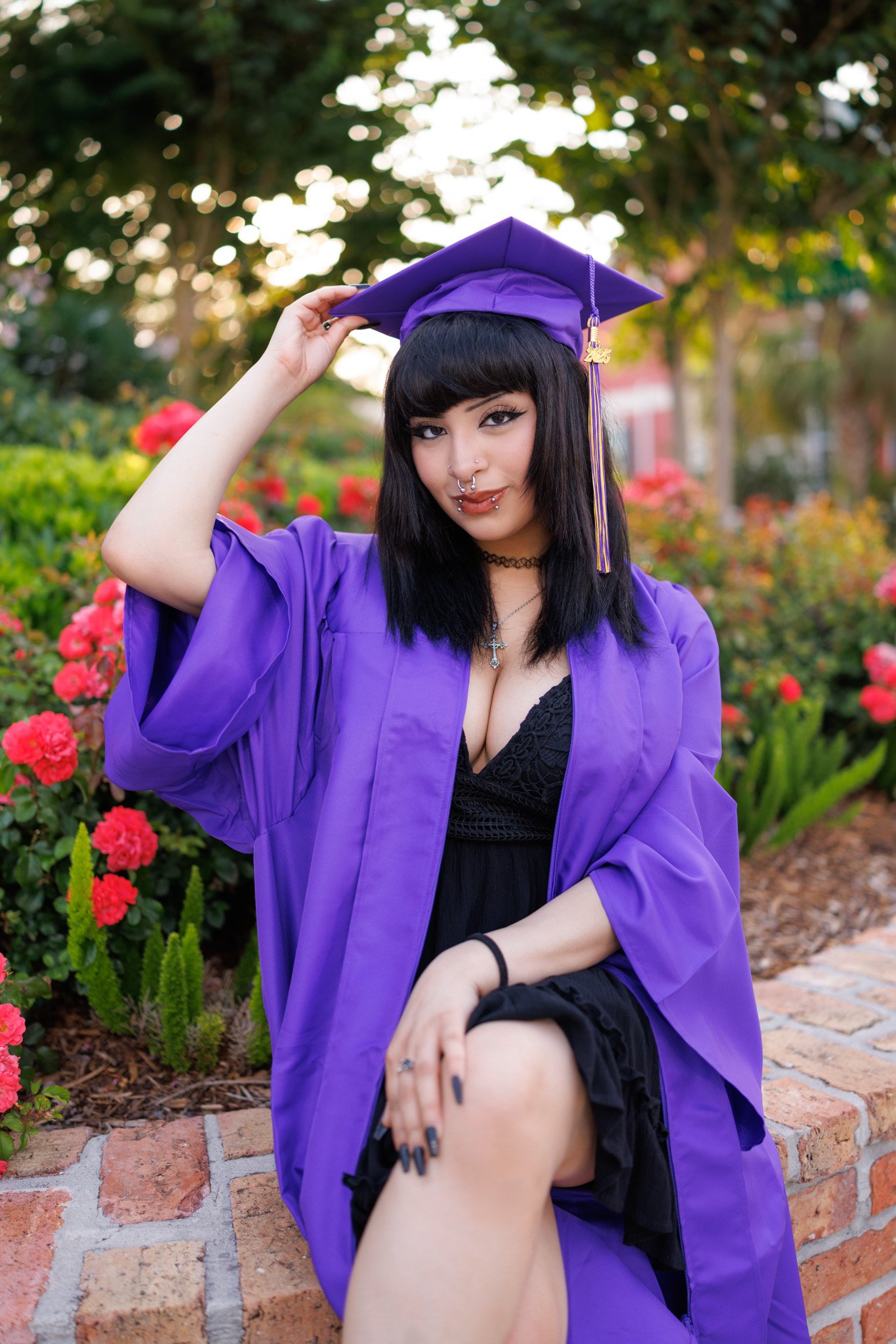 Young woman in graduation gown and cap, sitting on a brick ledge in a garden with pink flowers, looking at the camera, smiling, with black hair and tattoos.