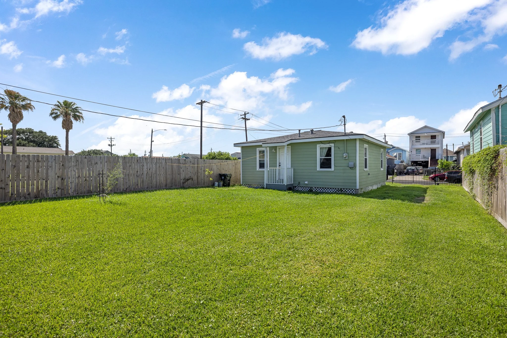 Backyard with green grass, wooden fence, and houses in the background under a blue sky with clouds.