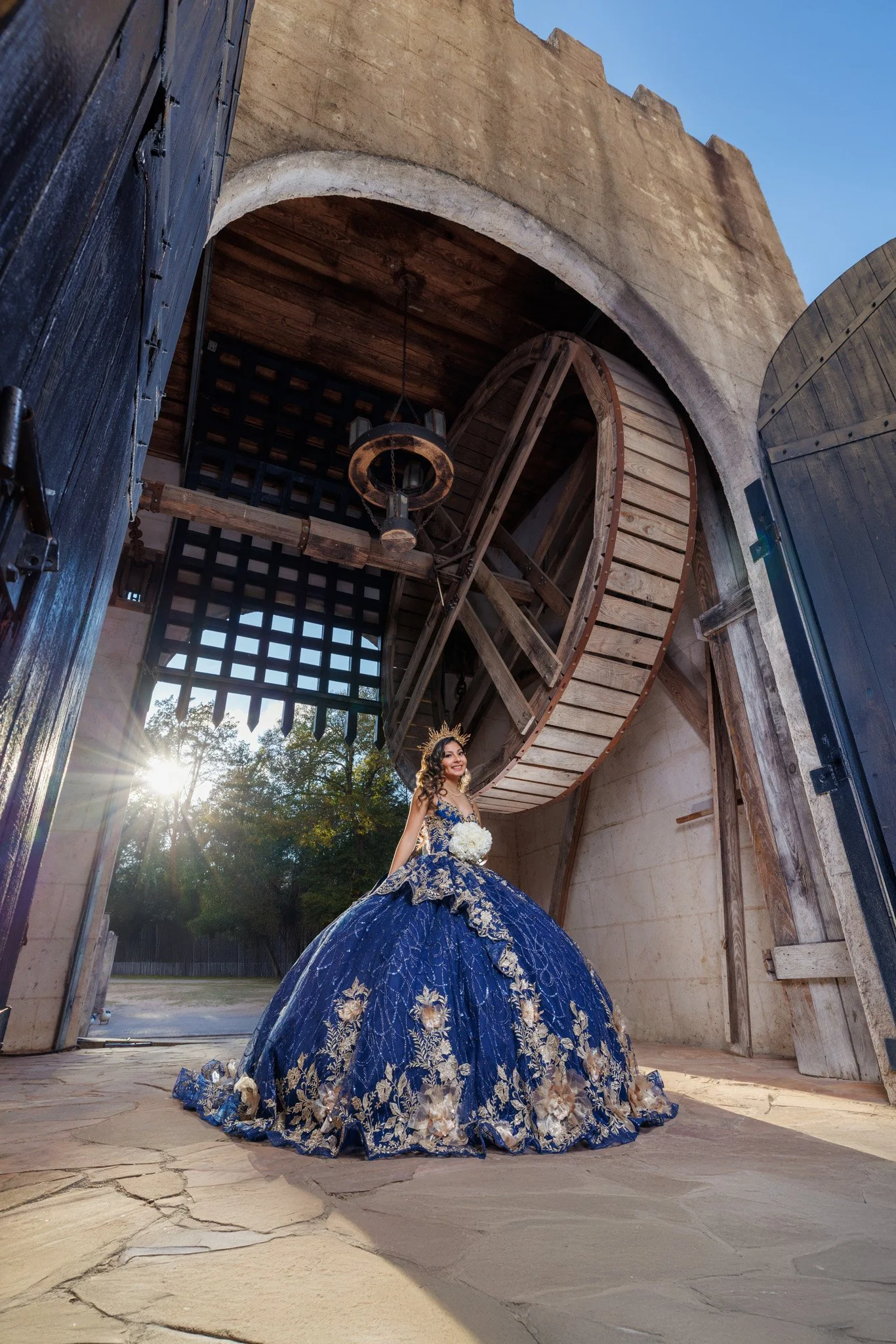 A woman in a blue and gold ball gown standing inside a vintage barn with open large wooden doors, holding a bouquet of white flowers, with sunlight shining through the trees outside.