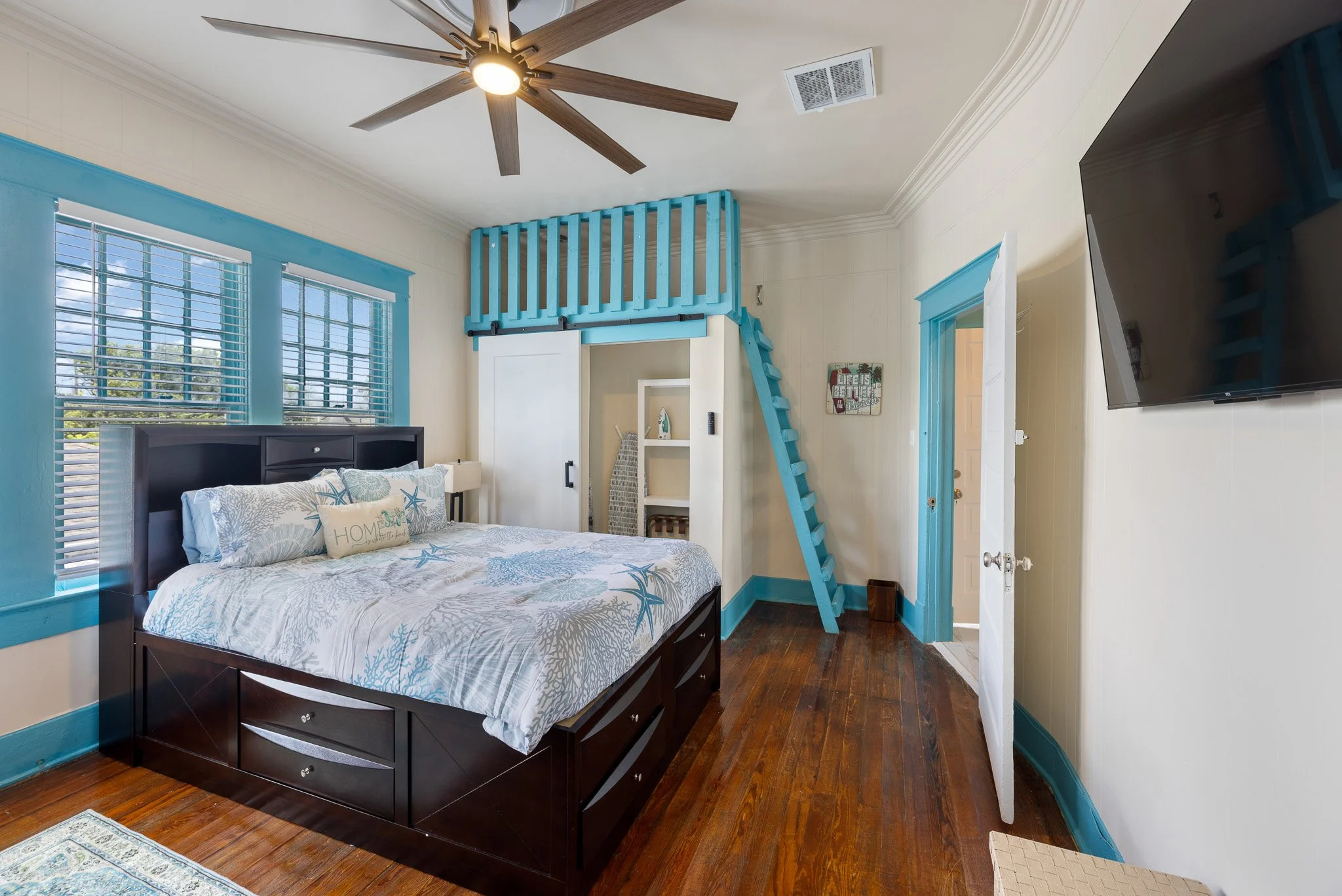 A bedroom with a black bed frame and ocean-themed bedding, two windows with blue trim, a ceiling fan, a wall-mounted TV, and a white and blue wooden loft with a ladder leading up to it.