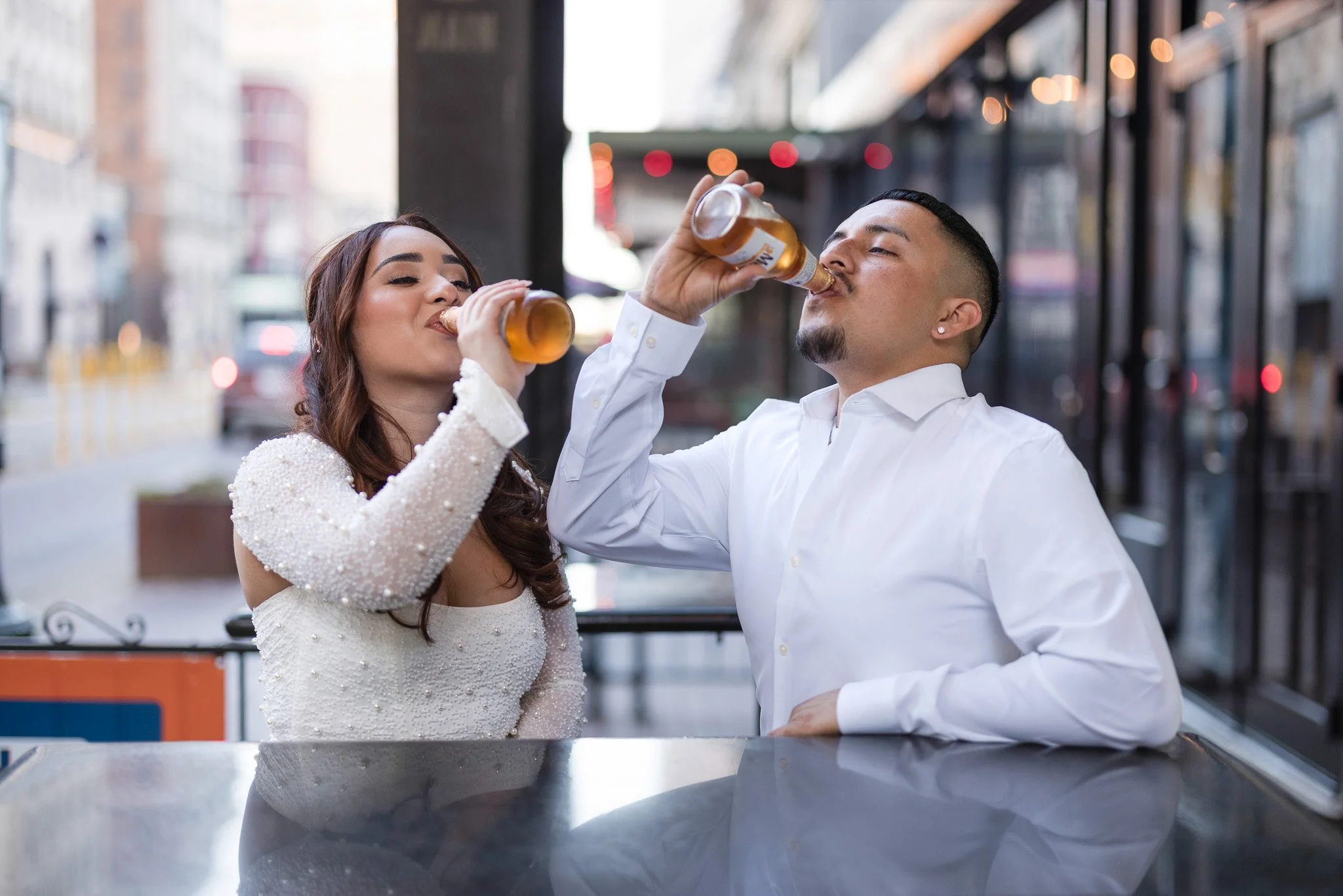 A woman and a man are sitting at a table outdoors, drinking beer in a city setting with buildings and lights in the background.