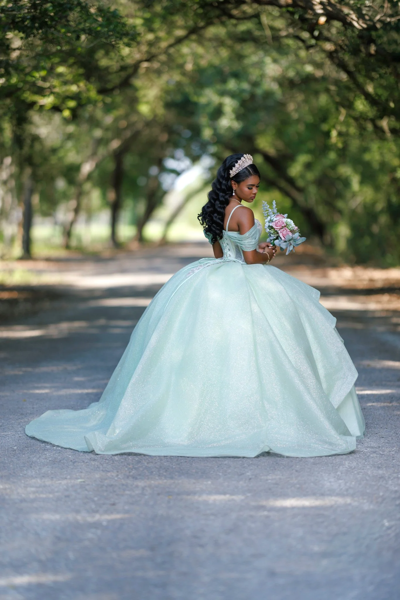 A woman in a light blue ball gown holding a bouquet of pink and white flowers in a shaded outdoor area with trees.