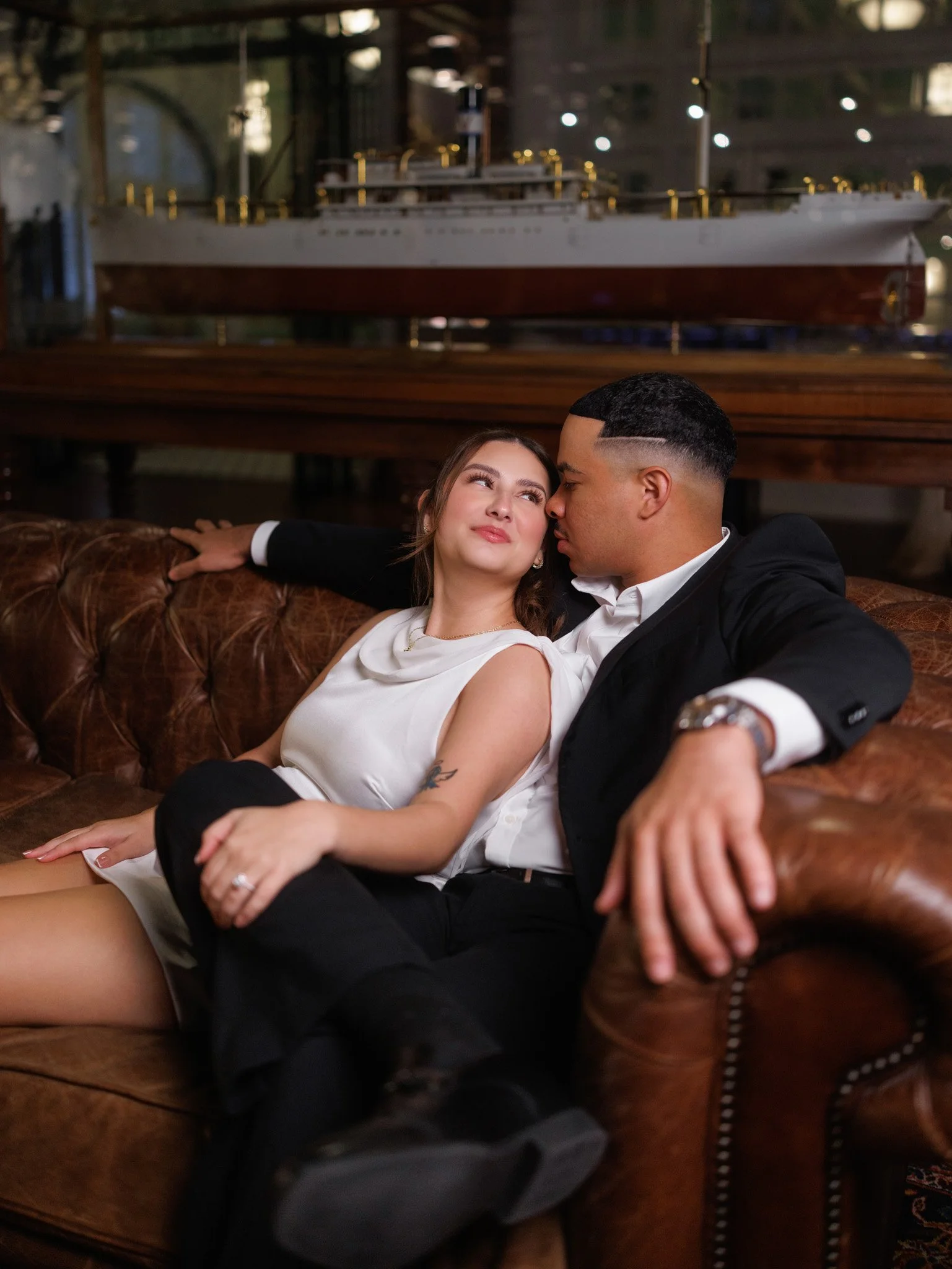 A couple sitting on a leather couch in a cozy, vintage-style room with a model ship in the background. The woman is wearing a white dress and the man is in a black suit with a white shirt.