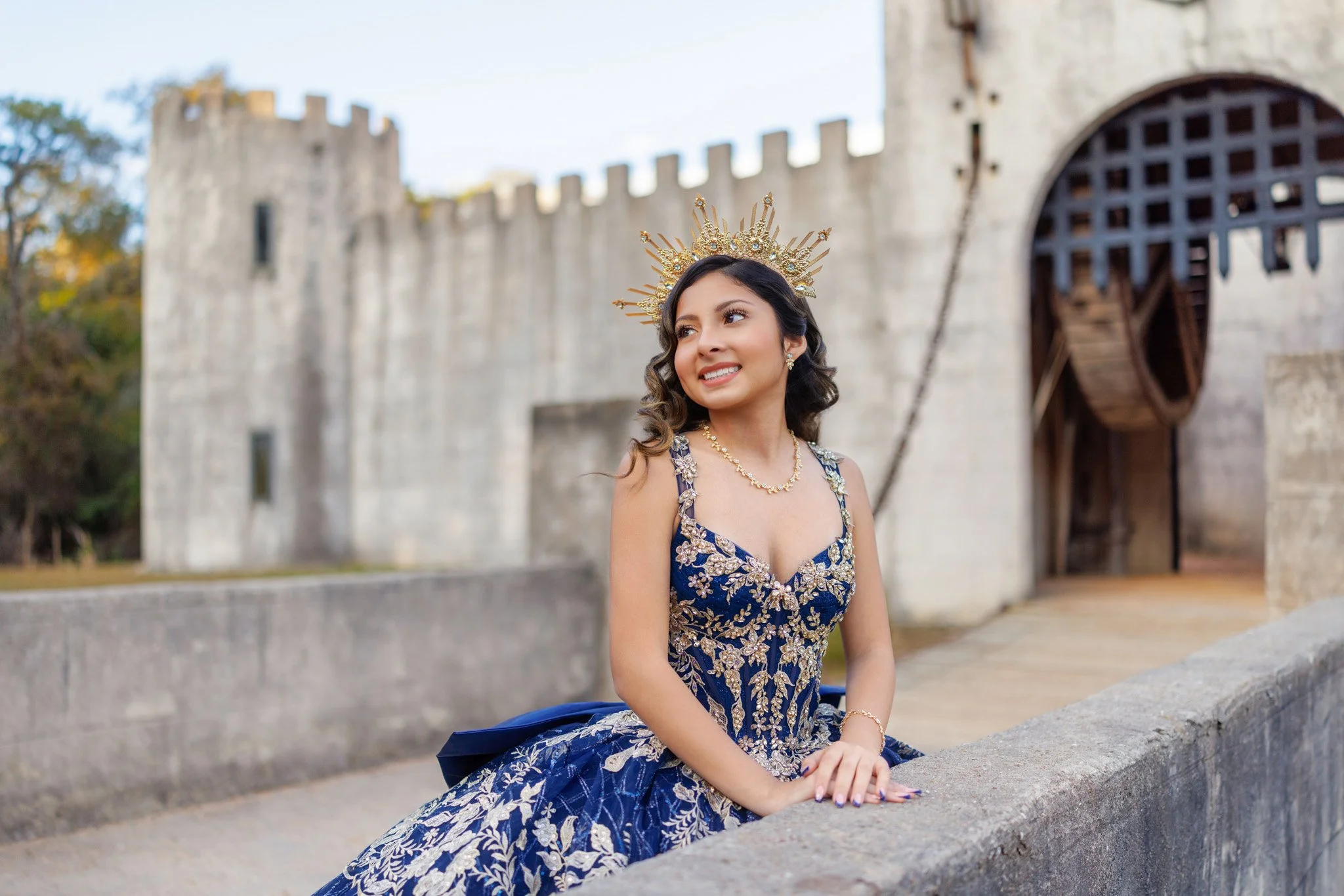 A woman in a royal blue dress with gold embroidery, sitting outdoors against a castle wall, wearing a gold crown and smiling.